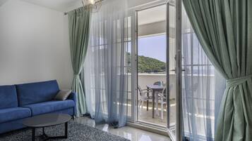 A room with a blue sofa, a small table, sheer curtains, and a view of a balcony with outdoor furniture.