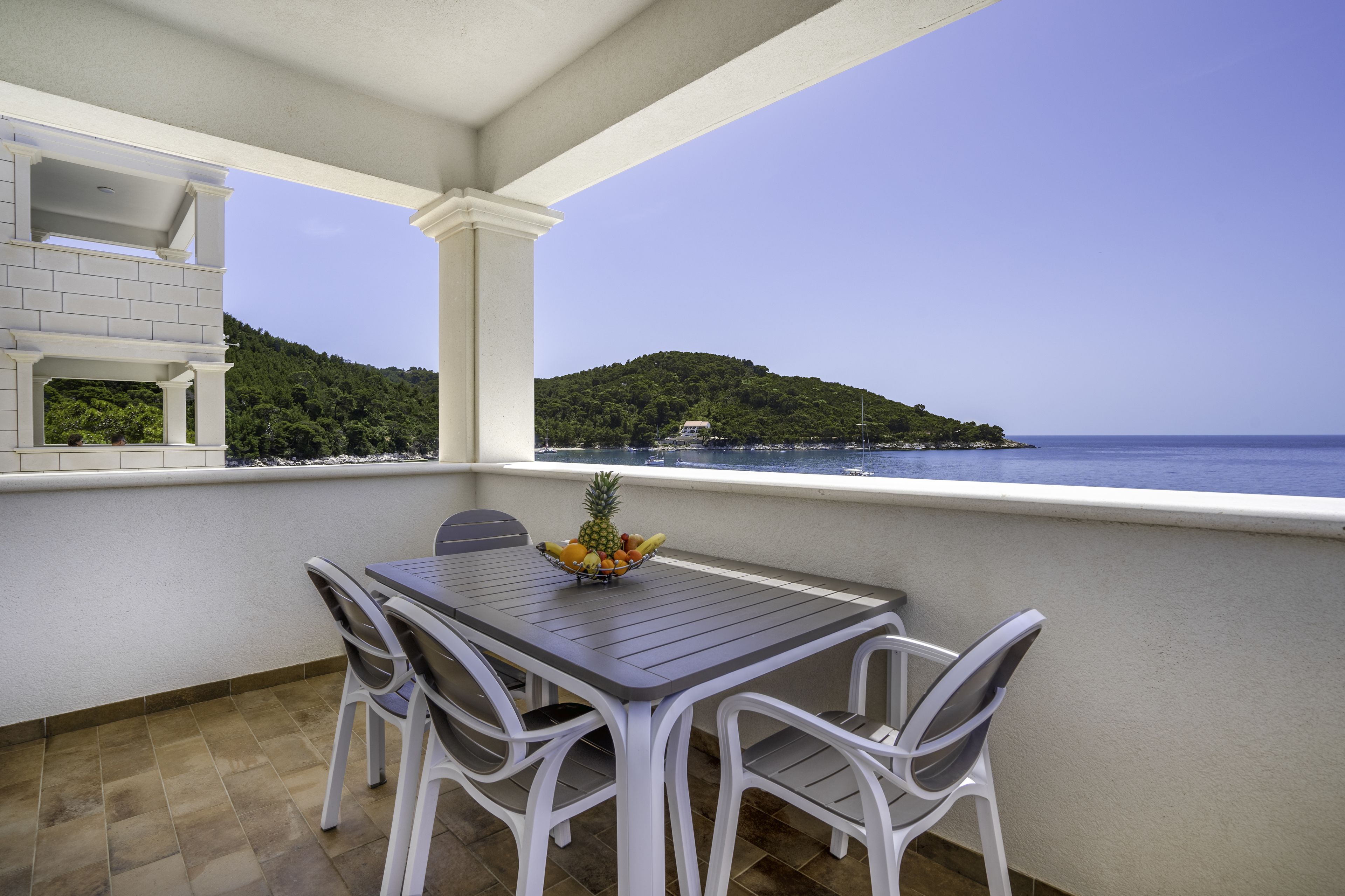 Un balcón con mesa y sillas, un frutero y vistas a una isla verde y al mar.