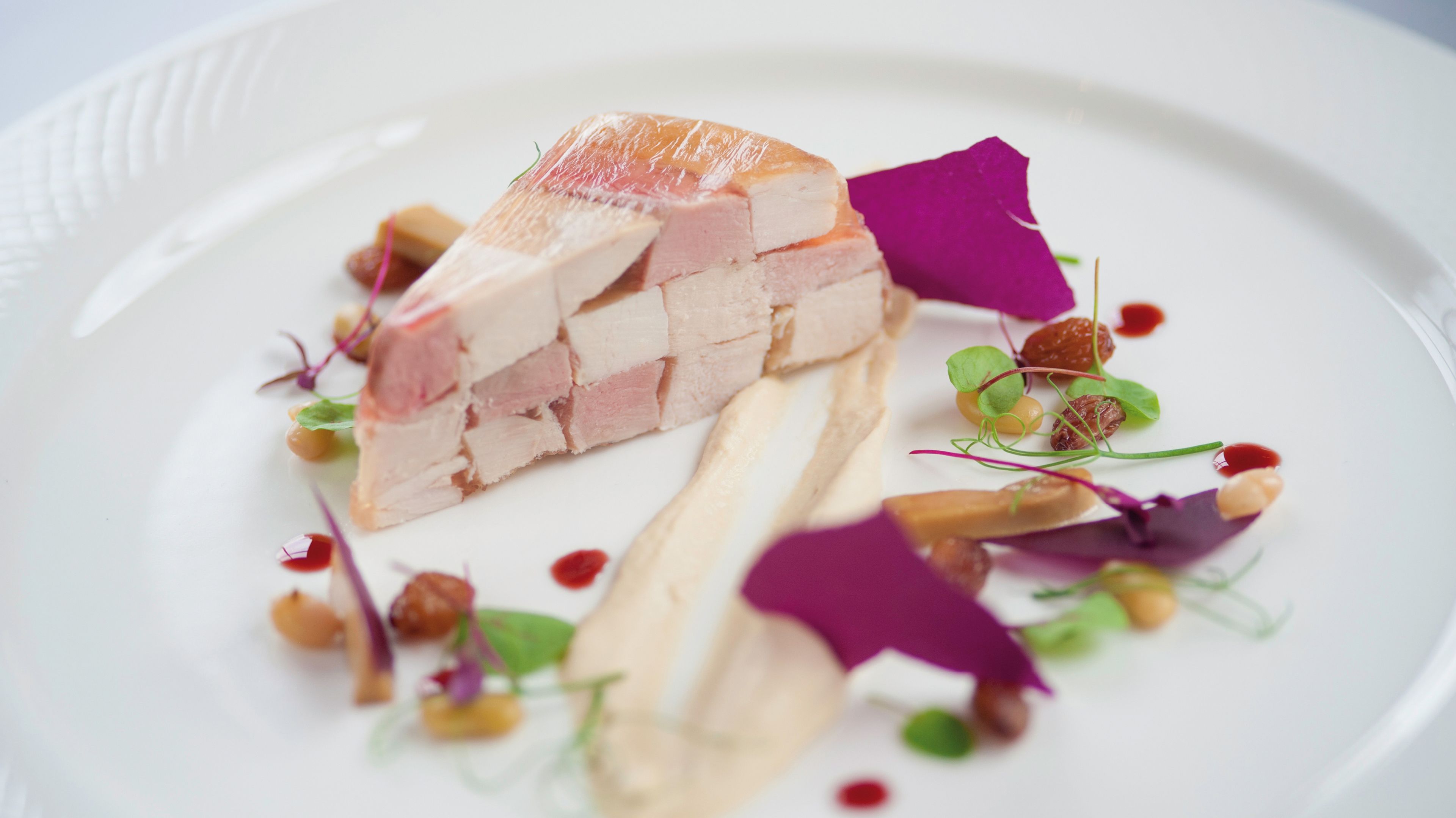 A white plate with a slice of meat, garnished with purple leaves and small green buds.