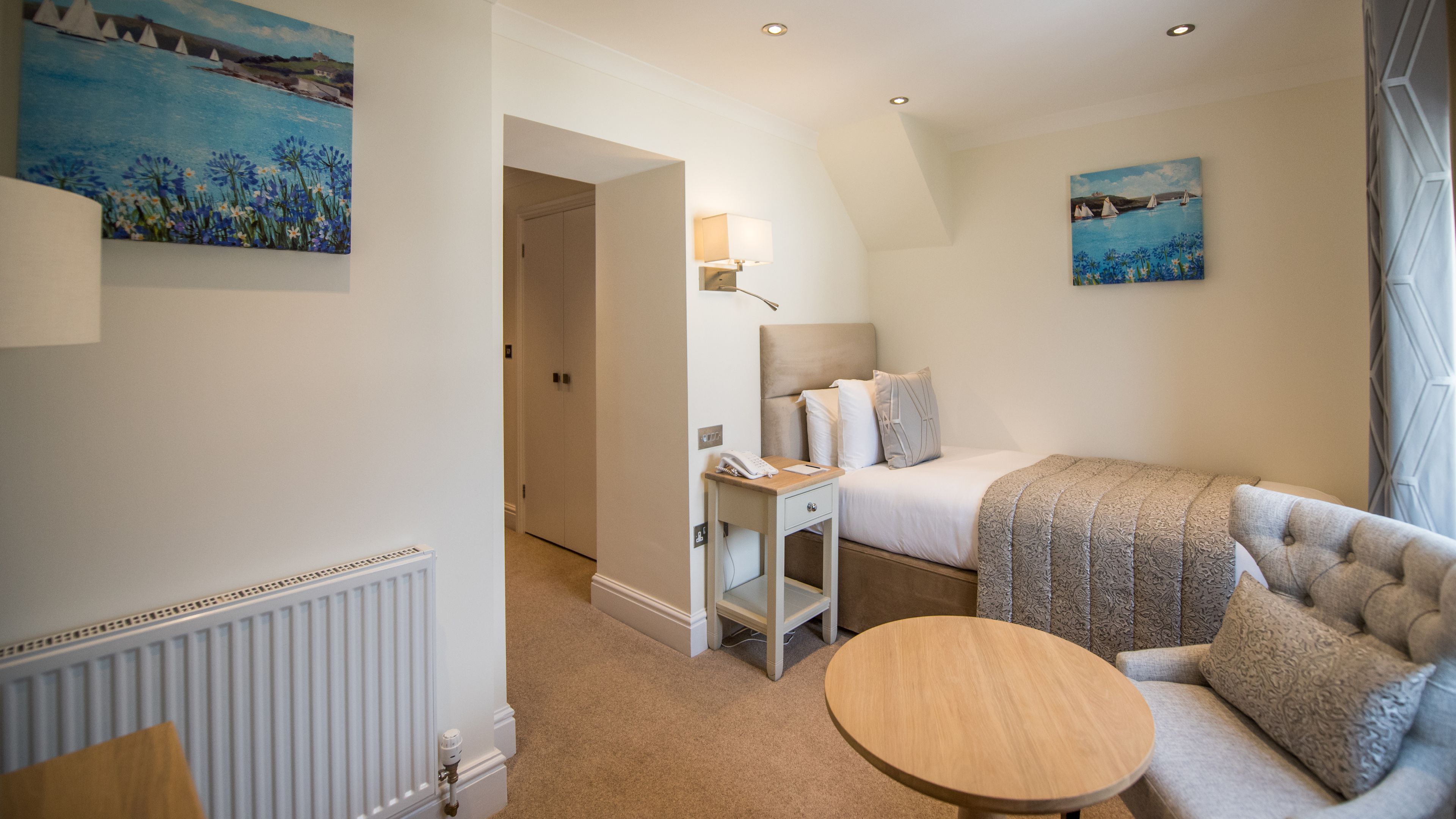 A hotel room with a bed, a bedside table with a lamp, a round wooden table, and two paintings on the walls.