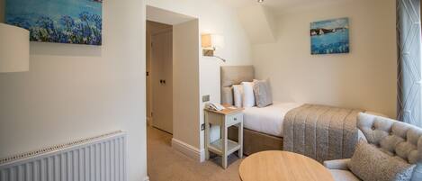 A hotel room with a bed, a bedside table with a lamp, a round wooden table, and two paintings on the walls.