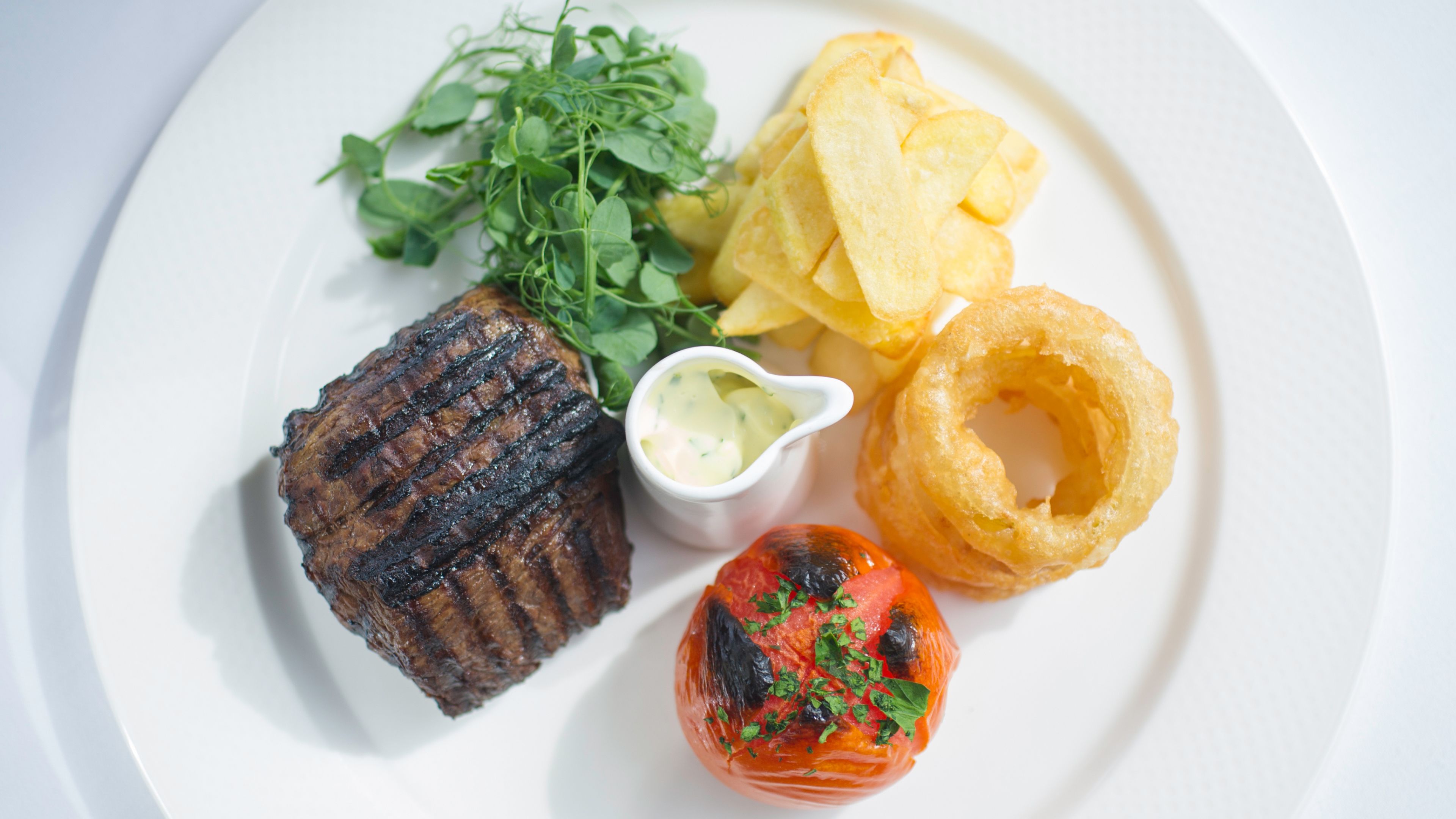 A plate with grilled steak, potato chips, onion rings, a tomato, and a small bowl of sauce.