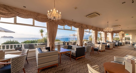 A spacious lounge area with multiple seating options, large windows offering ocean views, and elegant chandeliers.