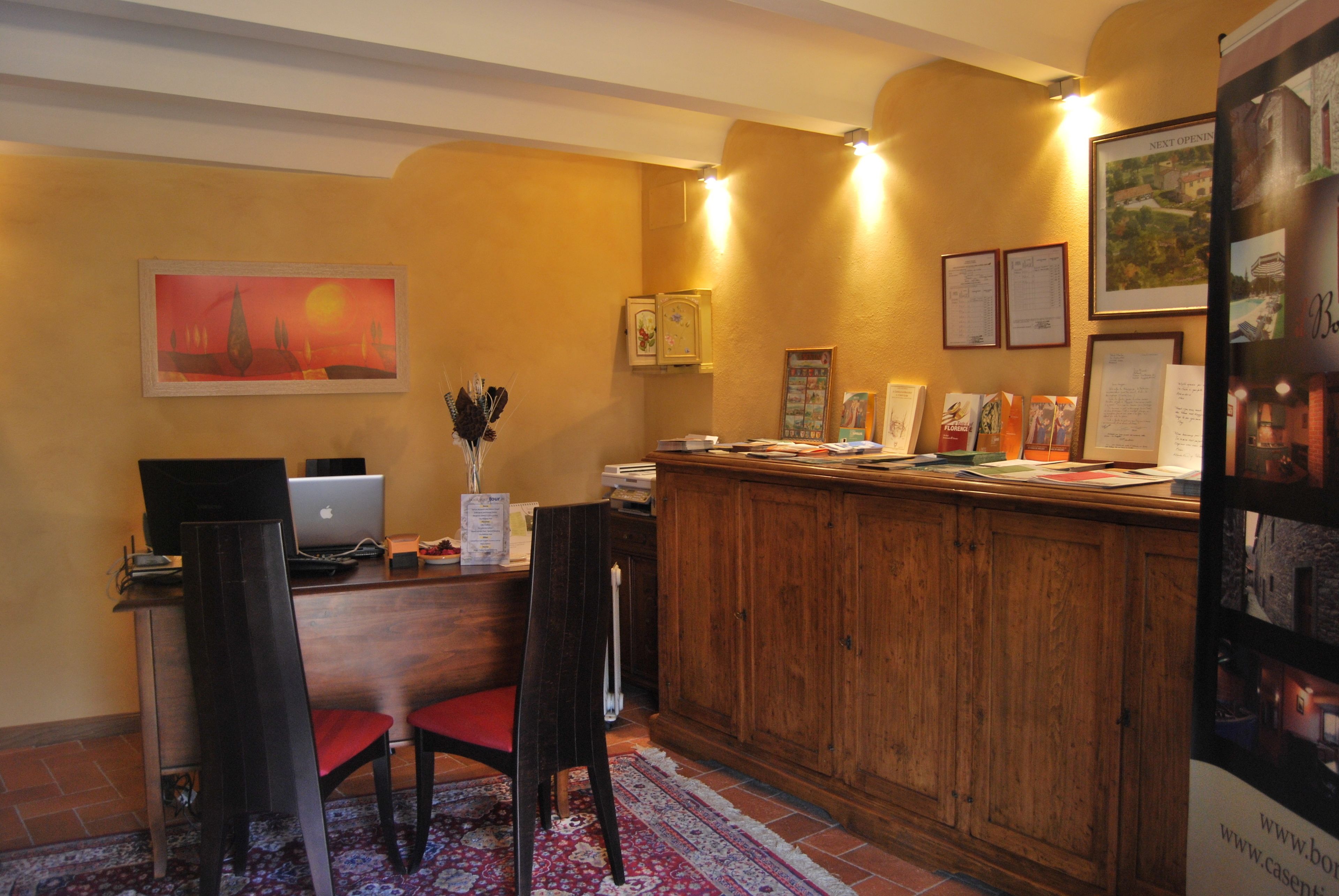 Un'area reception con una scrivania in legno, un portatile, un vaso con fiori e quadri appesi al muro.