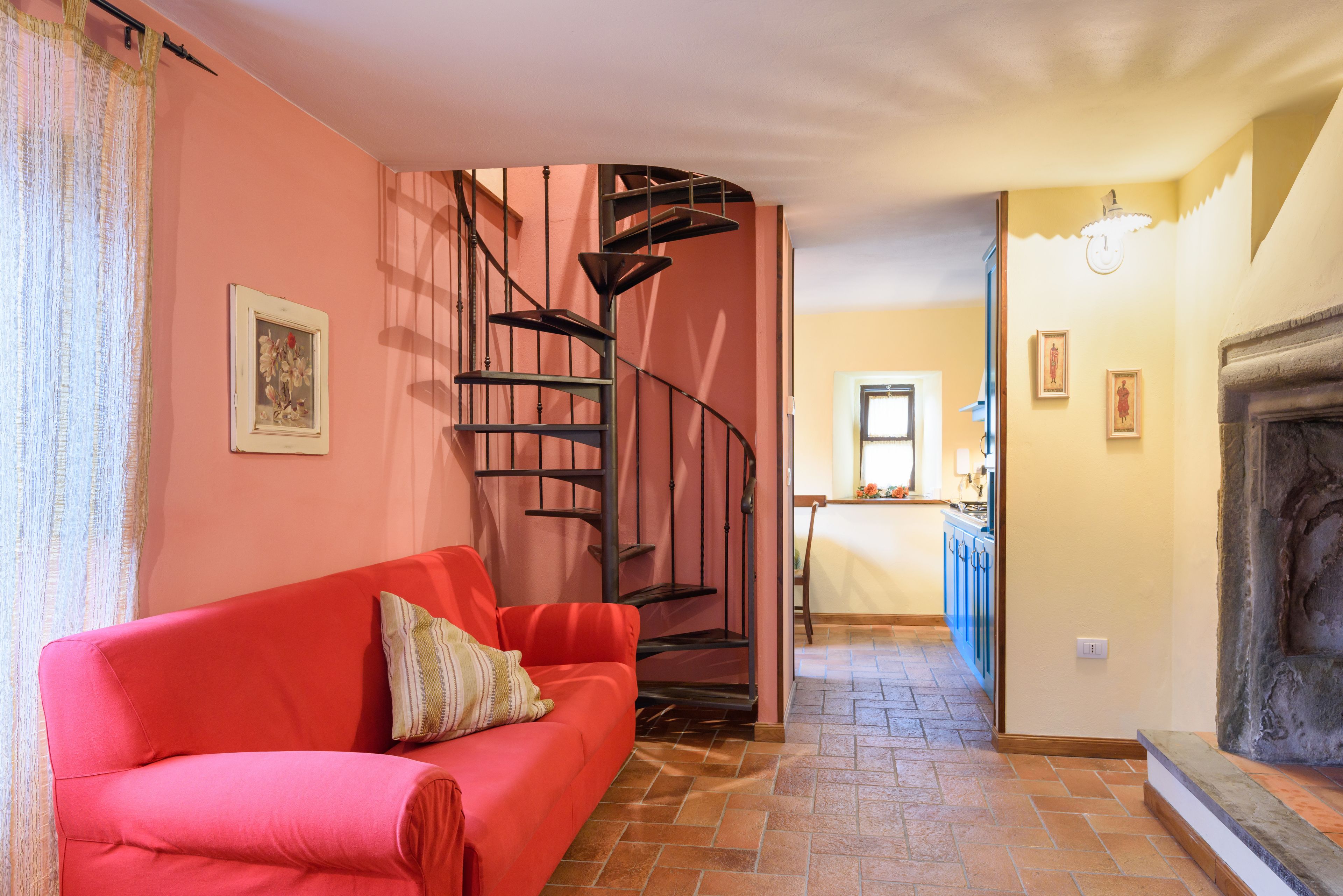 Una sala de estar con un sofá rojo, una escalera de caracol, una cocina al fondo y una chimenea.