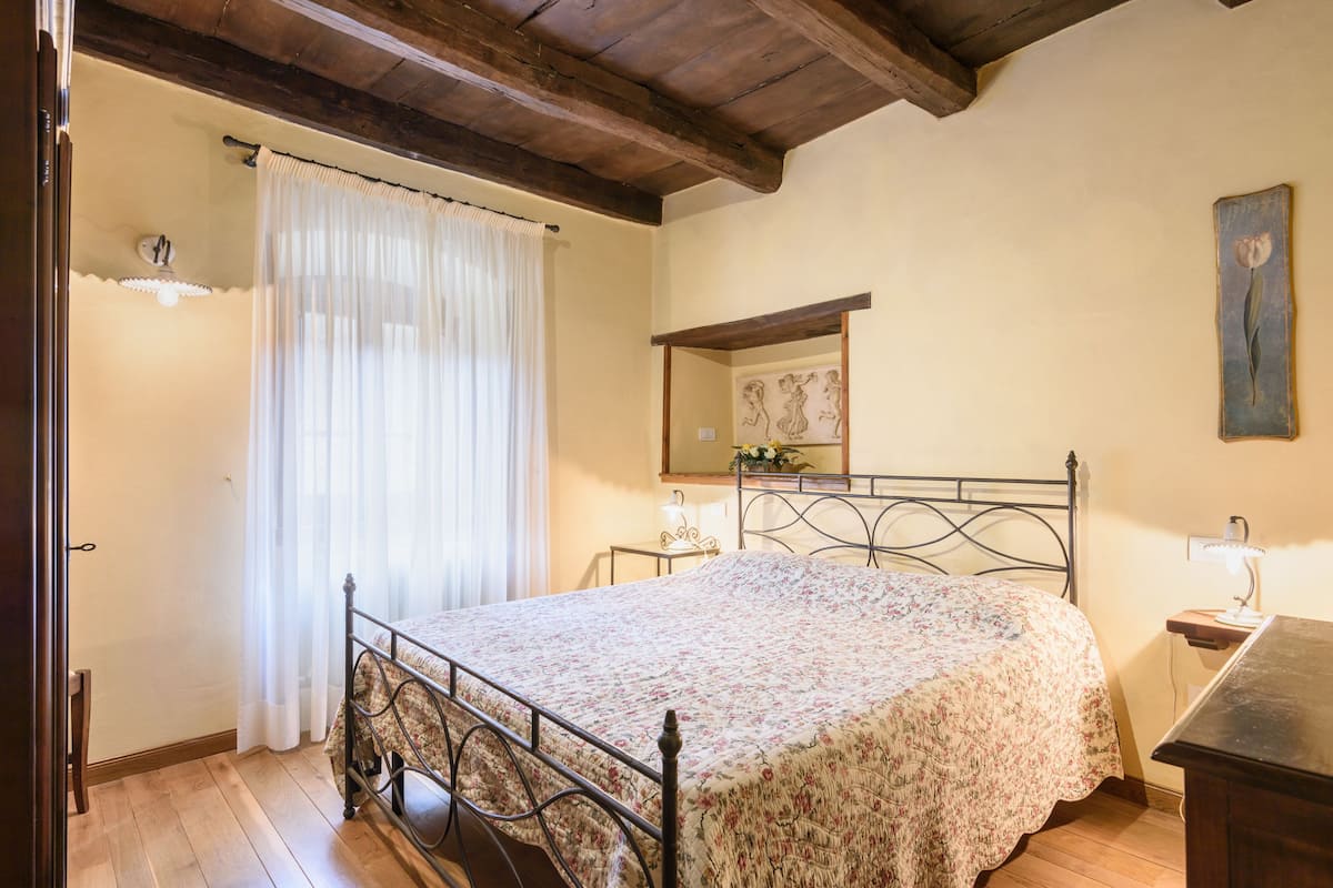 A bedroom with a bed, wooden floor, a ceiling with exposed beams, a bedside table with a lamp, and a wall-mounted mirror.