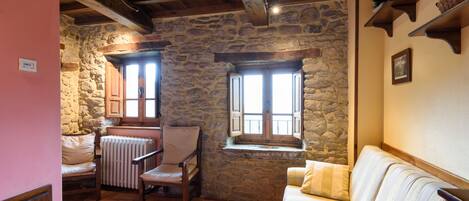 Una habitación acogedora con pared de piedra, vigas de madera, un banco acolchado, un sofá y una ventana con contraventanas.