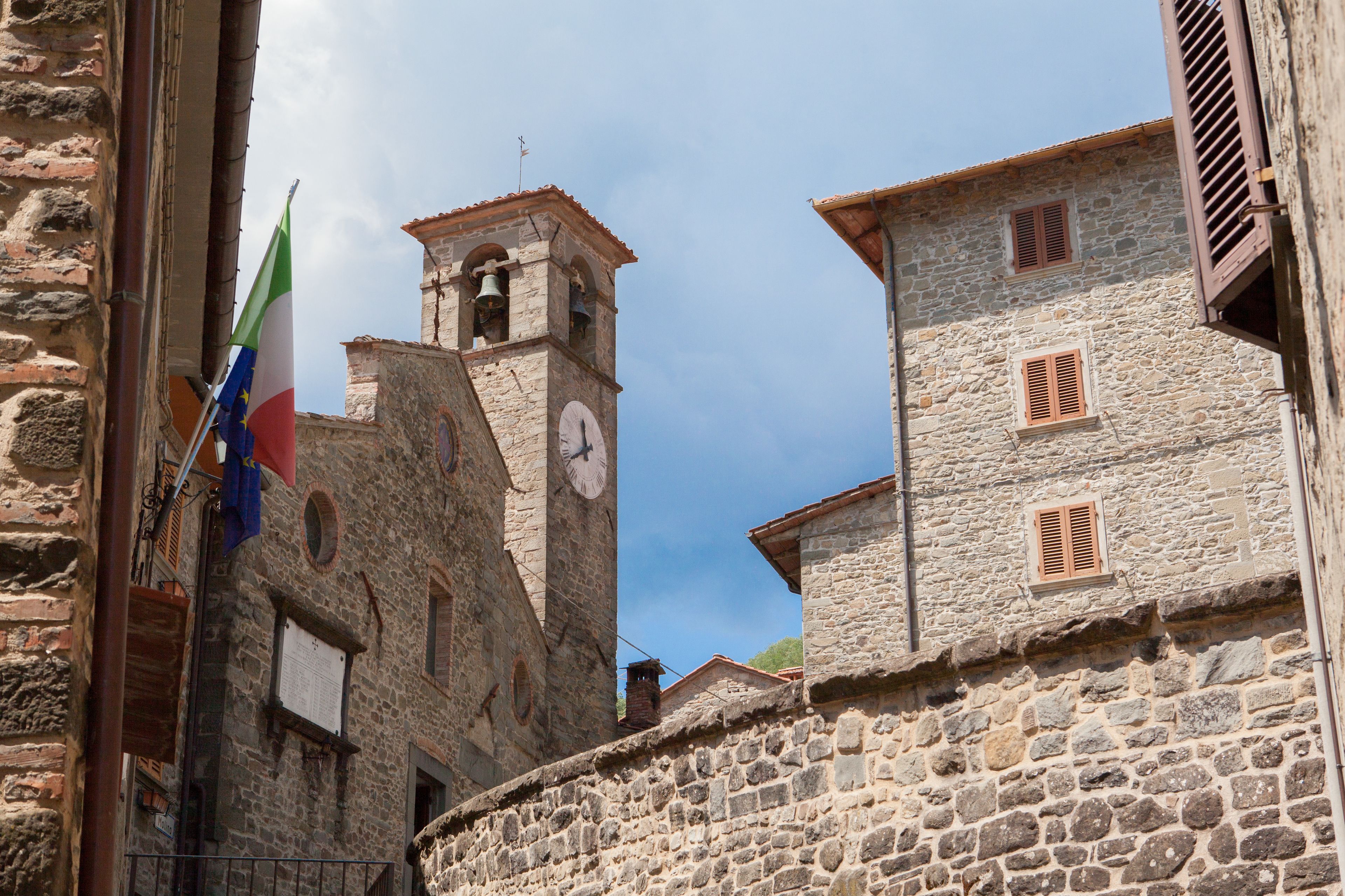 Un edificio de piedra con una torre de reloj, contraventanas de madera y una bandera.