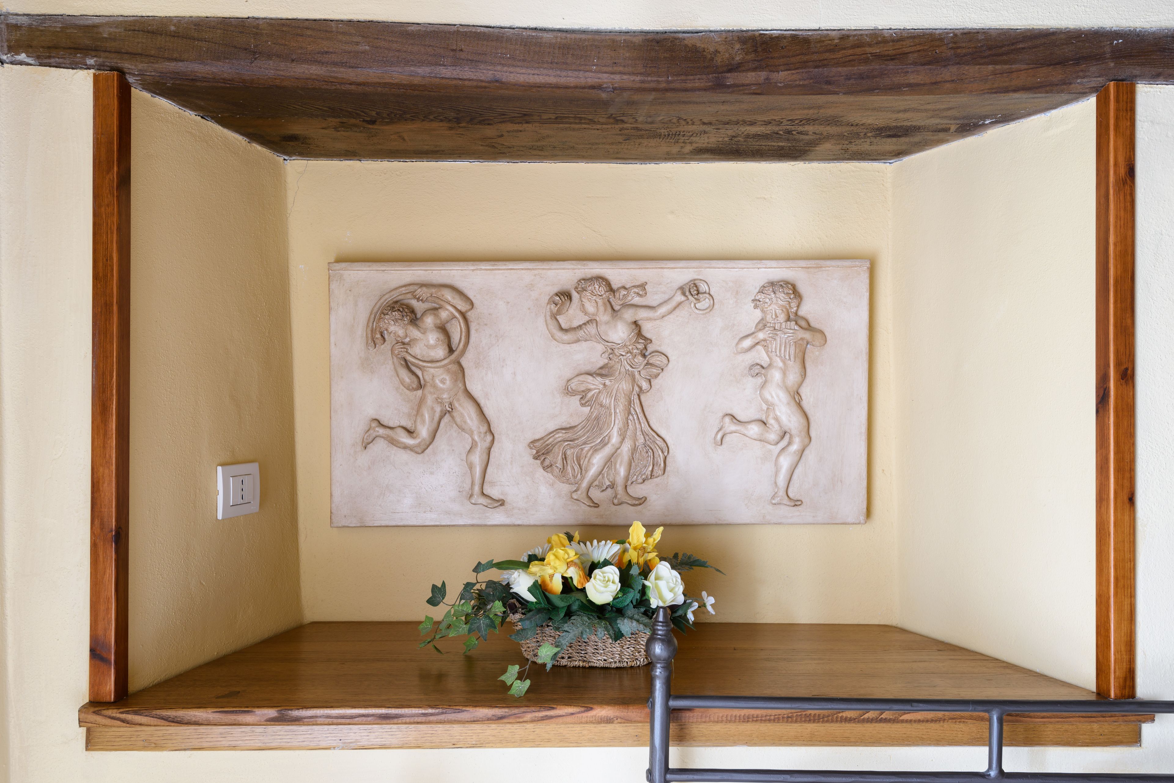 Una habitación con un banco de madera, una obra de arte en la pared que representa tres figuras y un arreglo floral sobre el banco.