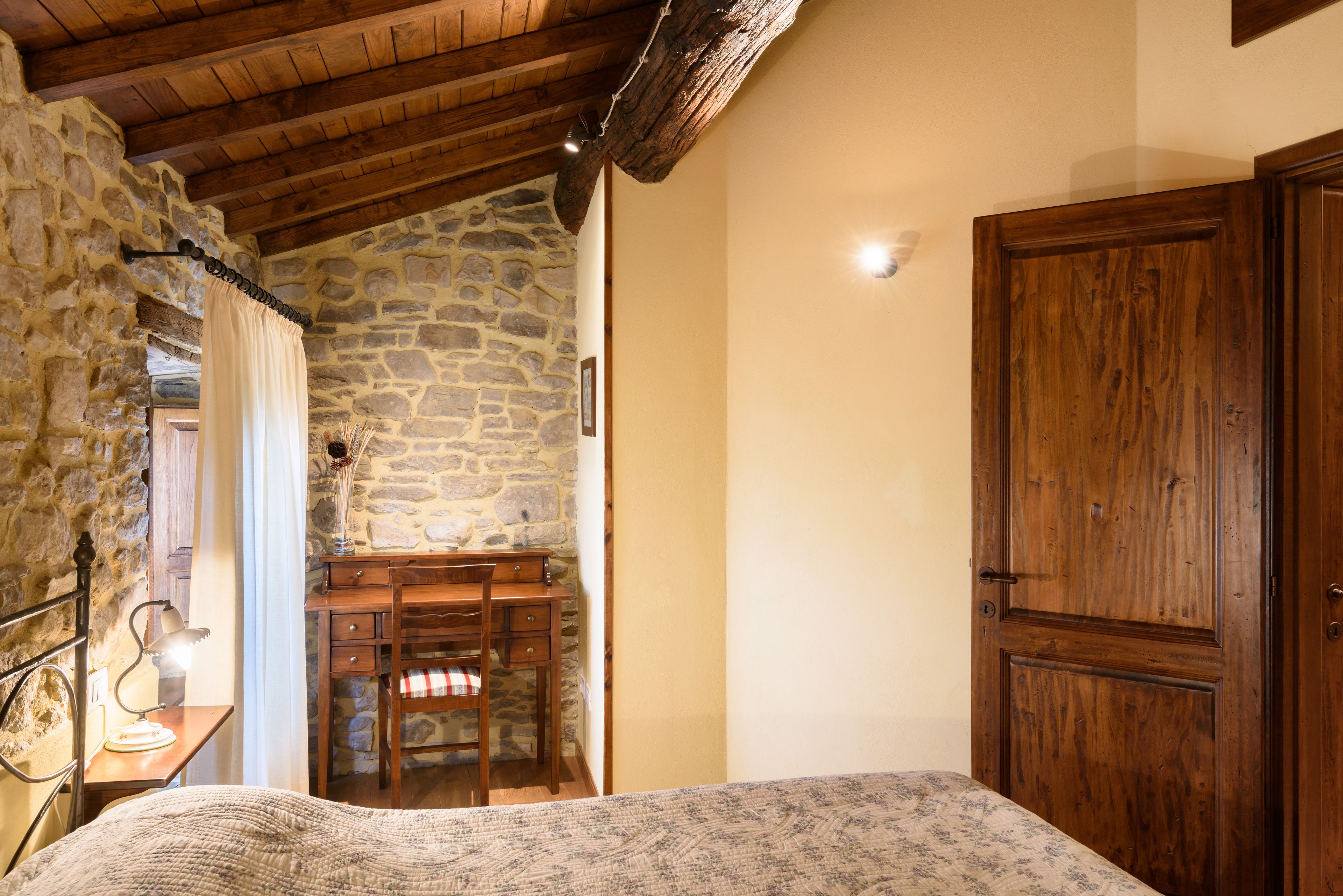 A bedroom with a stone wall, wooden ceiling beams, a bed, a desk, and a wardrobe.