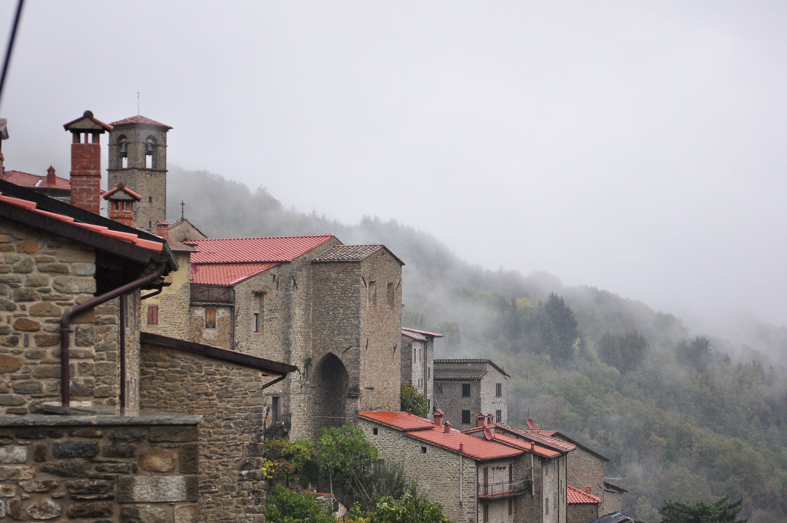 A stone building with red-tiled roofs and a bell tower, surrounded by misty hills.