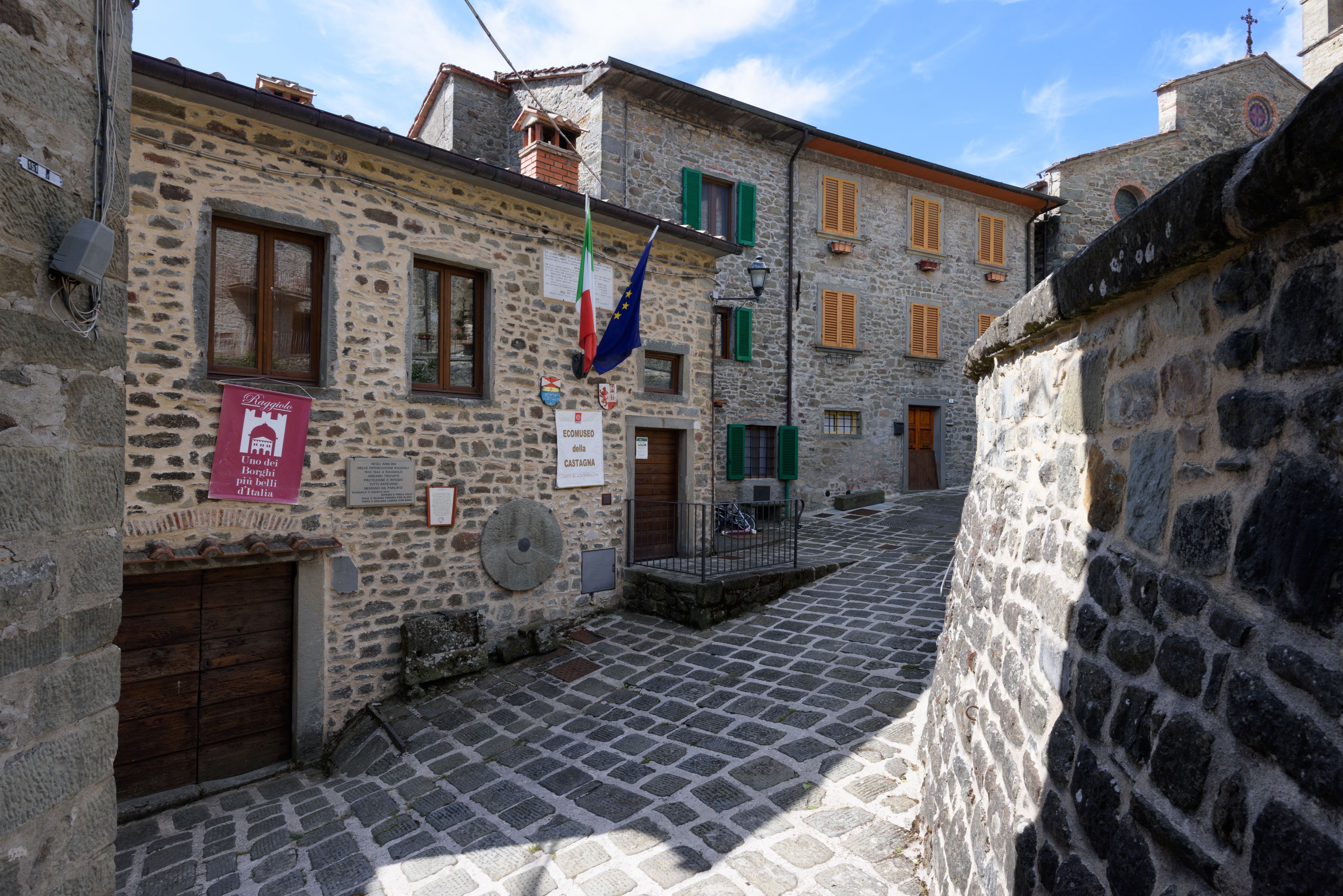 A narrow cobblestone street with stone buildings, a sign for a hotel, and a flag.