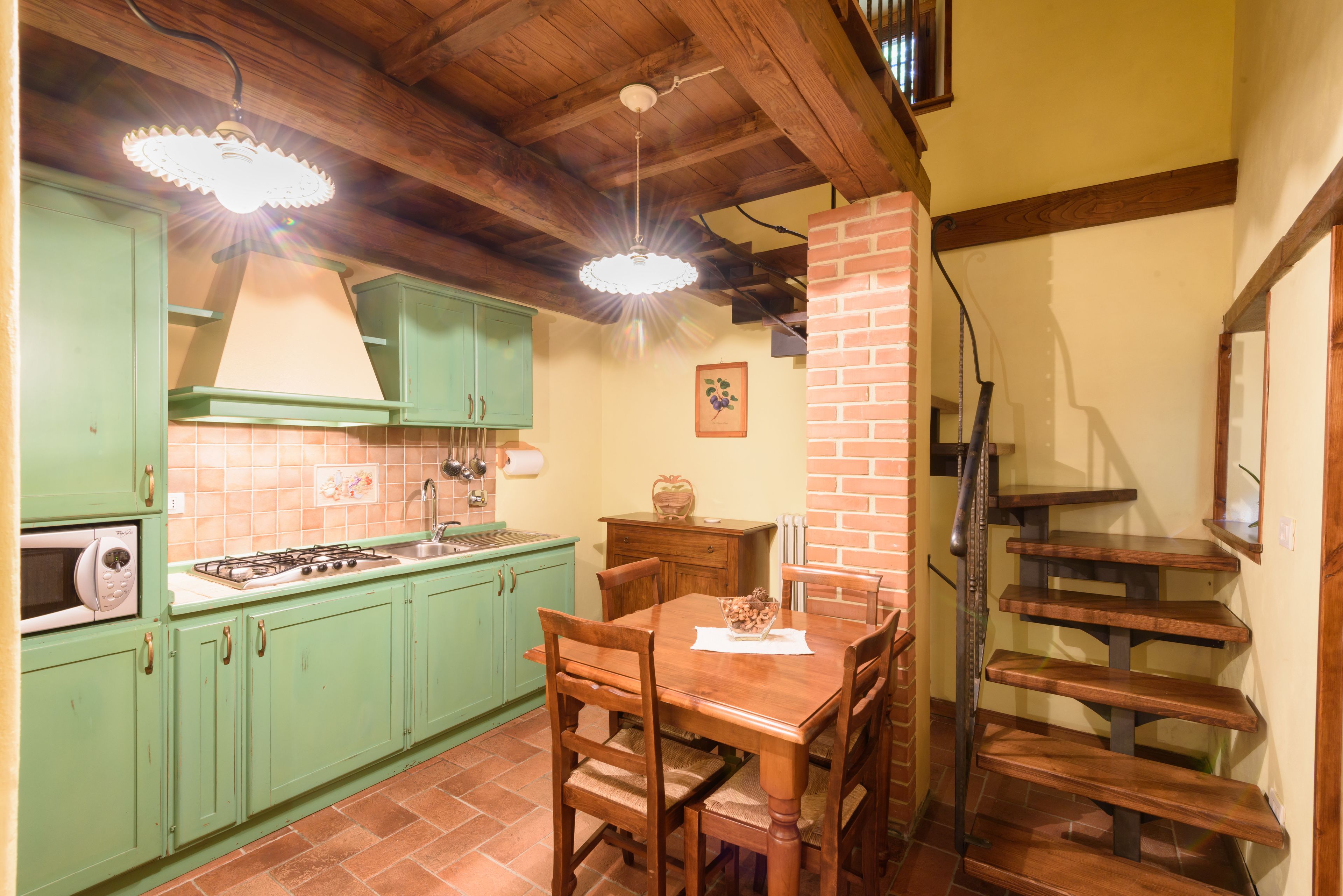 Une cuisine avec des meubles de rangement verts, une table à manger en bois et une cheminée en briques.