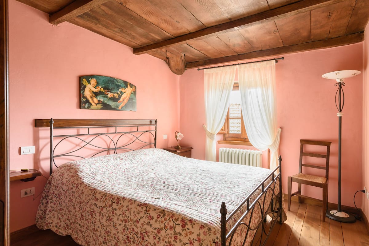 A bedroom with a bed, a nightstand, a chair, a floor lamp, a painting, and a window with curtains.