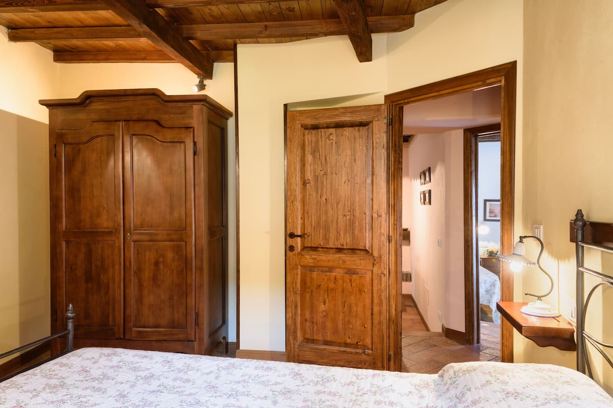 A bedroom with a wooden wardrobe, a bed with a floral bedspread, and a nightstand with a lamp.