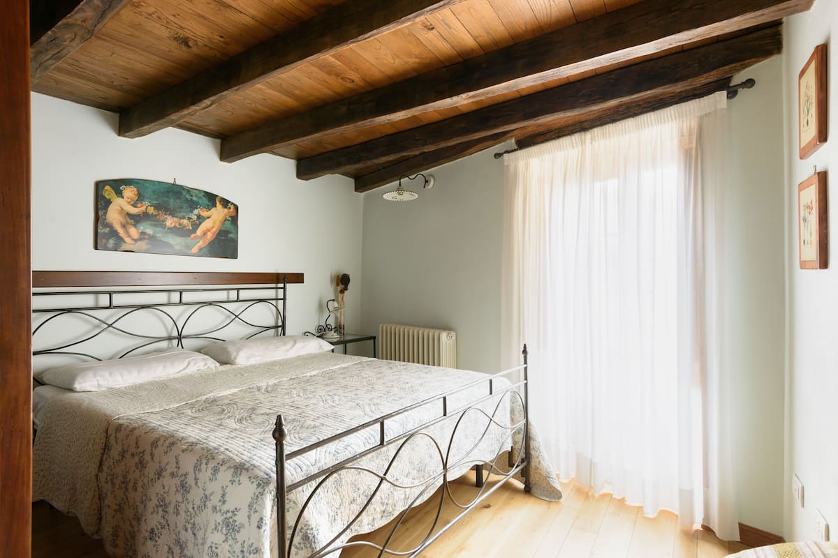 A bedroom with a wooden ceiling, a bed with a metal frame, a radiator, and a painting on the wall.