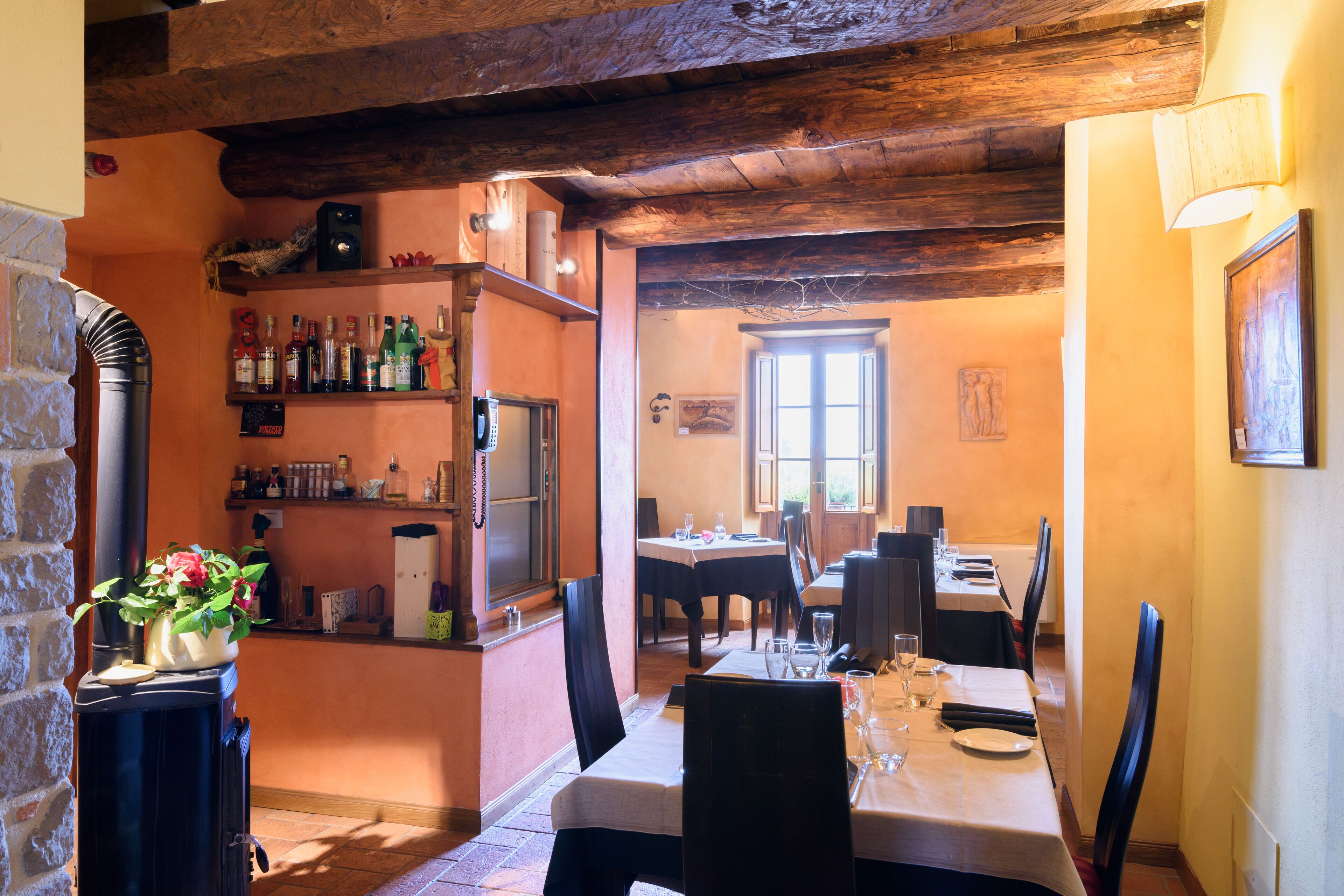 A dining area with wooden beams, a fireplace, and a bar with bottles. Tables are set for dining.