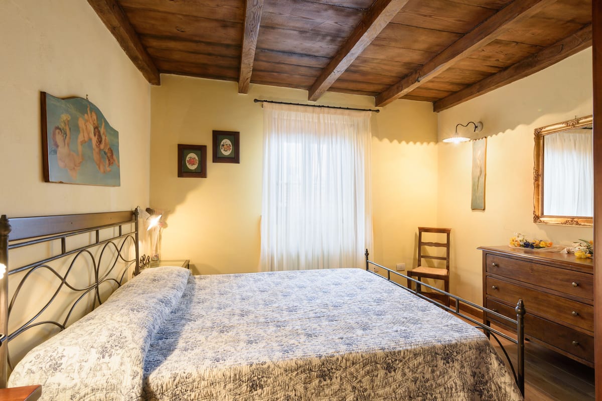 A bedroom with a bed, a wooden headboard, a dresser, a chair, a window with curtains, and a painting on the wall.