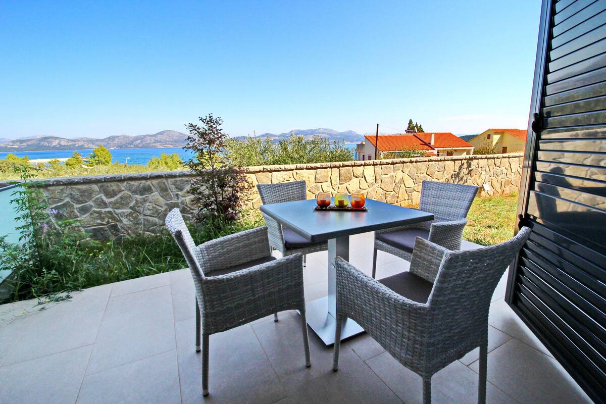 A patio with a table and four wicker chairs, overlooking a body of water and distant mountains.