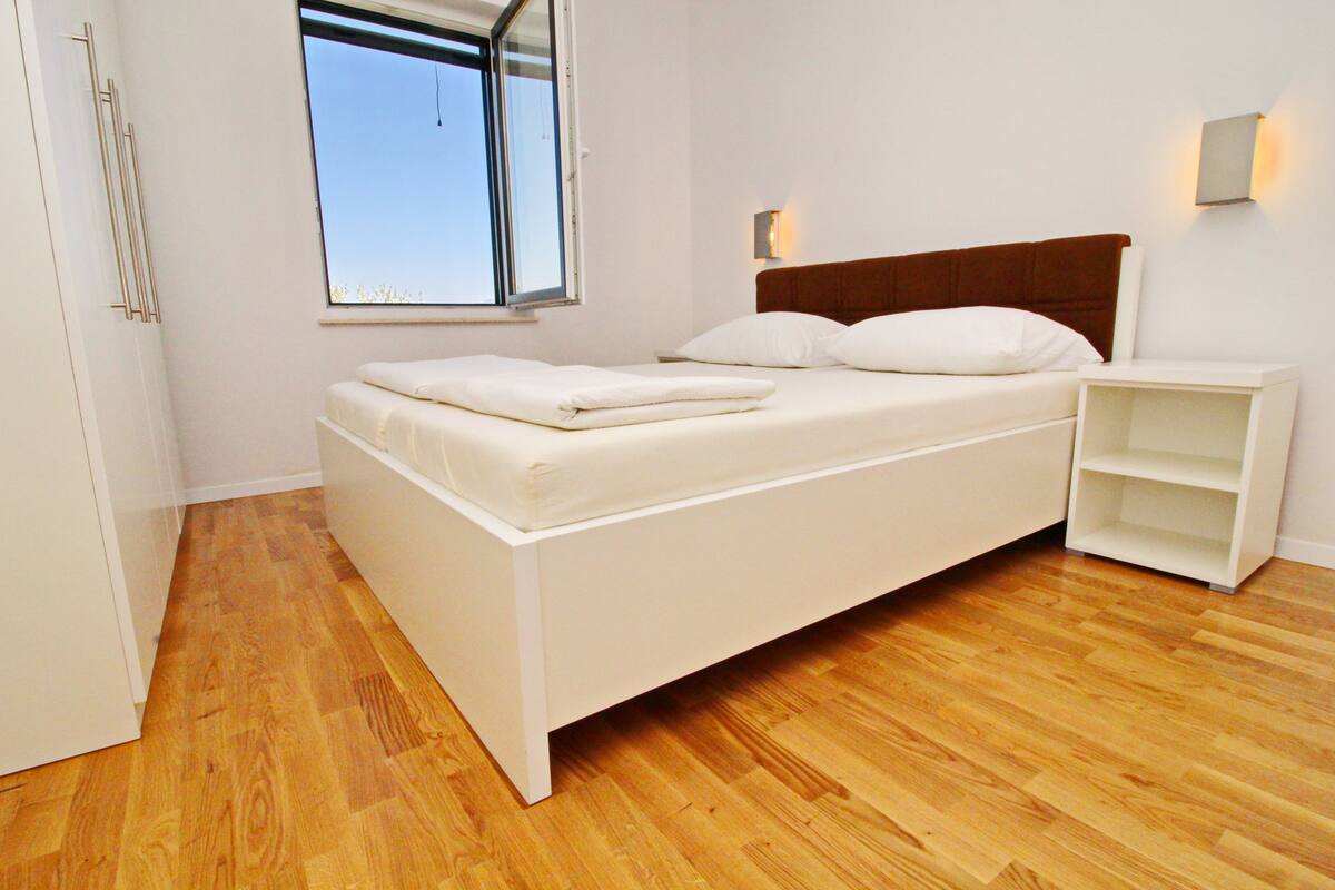 A bedroom with a bed, a nightstand, a window, and a wooden floor.