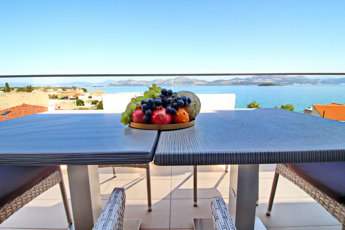 A dining table with a fruit bowl on it, overlooking a scenic view of the sea and mountains.