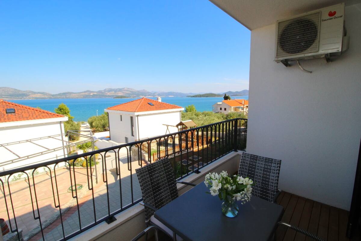 A balcony with a table and chairs, overlooking a coastal view with houses and a clear sky.