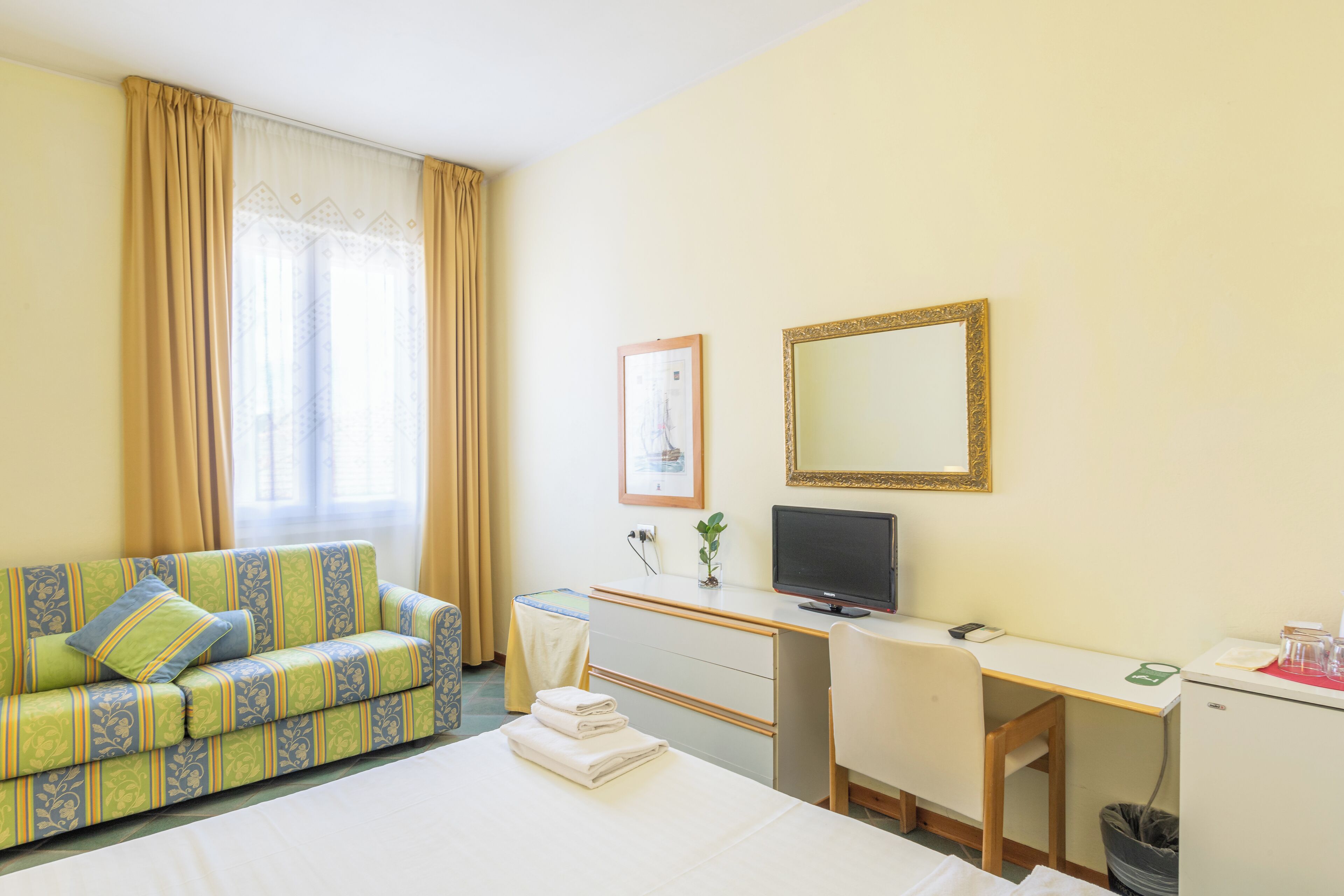 A hotel room with a bed, a sofa, a desk, a television, and a small trash bin.