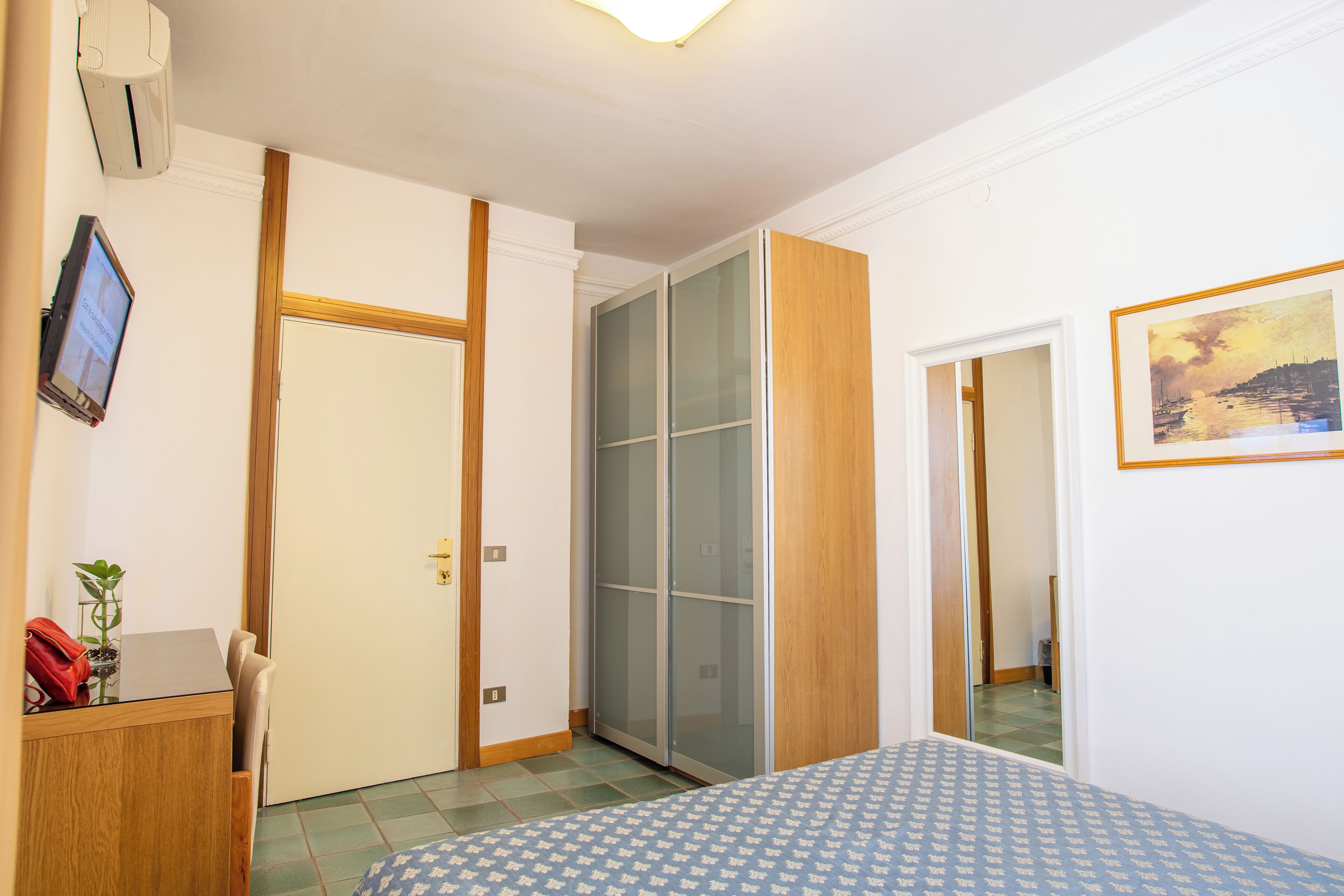 A bedroom with a bed, a desk, a chair, a wardrobe, and a painting on the wall.