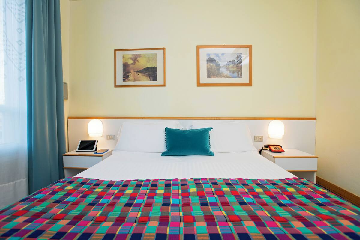 A hotel room with a large bed, a colorful quilt, two bedside tables with lamps, and two framed pictures on the wall.