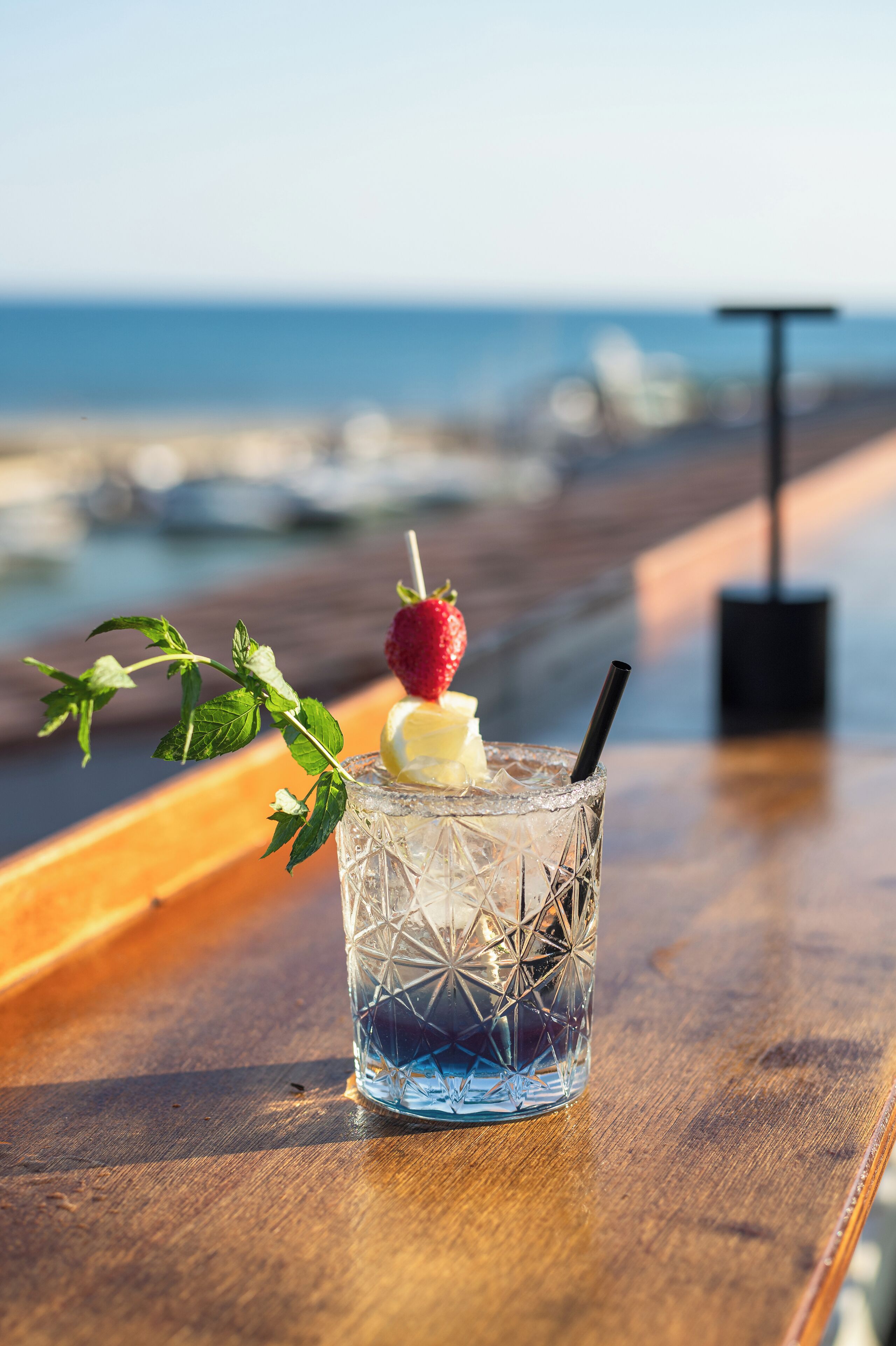 A glass with a strawberry and mint garnish on a wooden table by the beach.