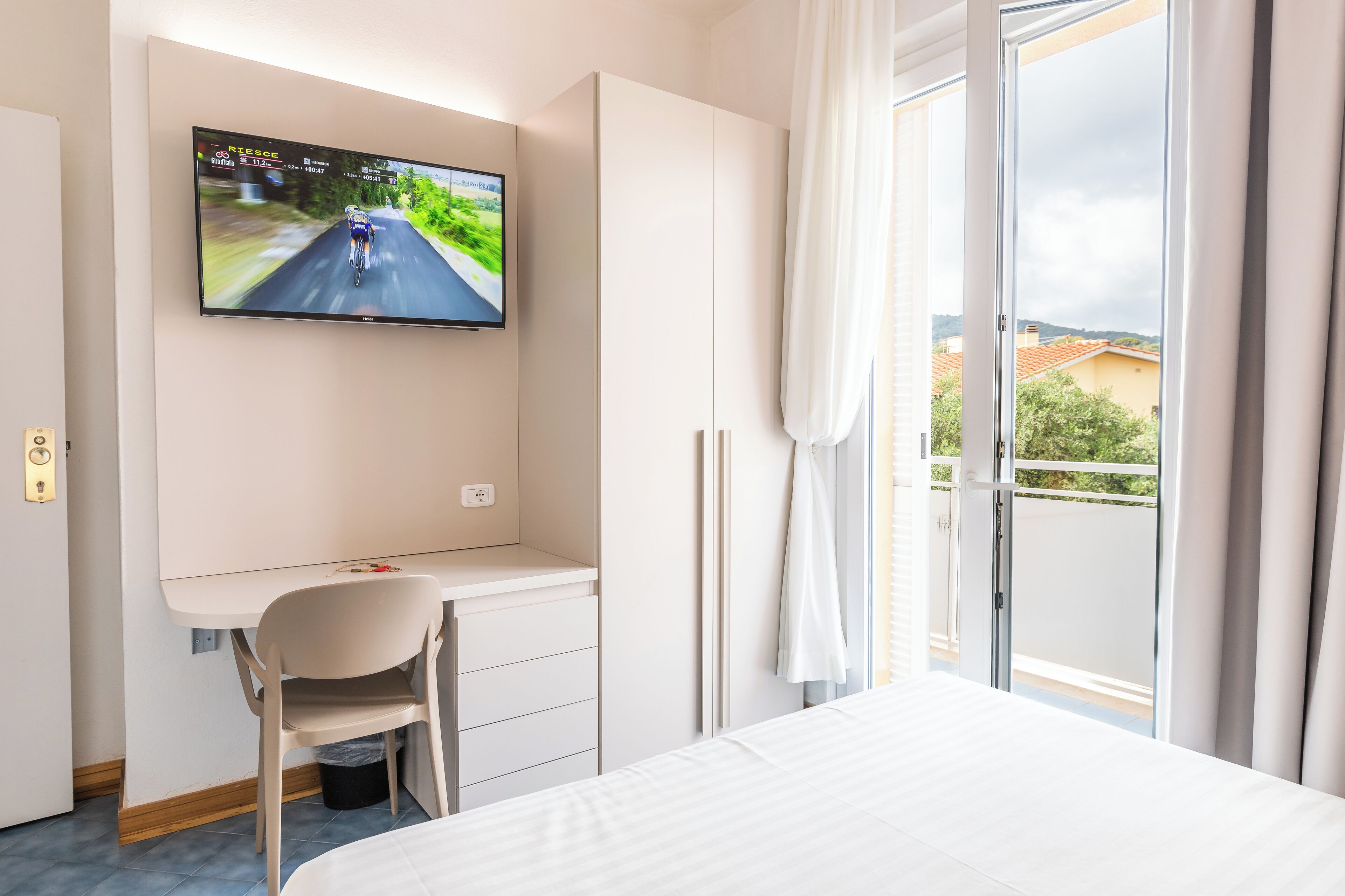 A hotel room with a bed, a desk with a chair, a TV mounted on the wall, and a balcony with curtains.