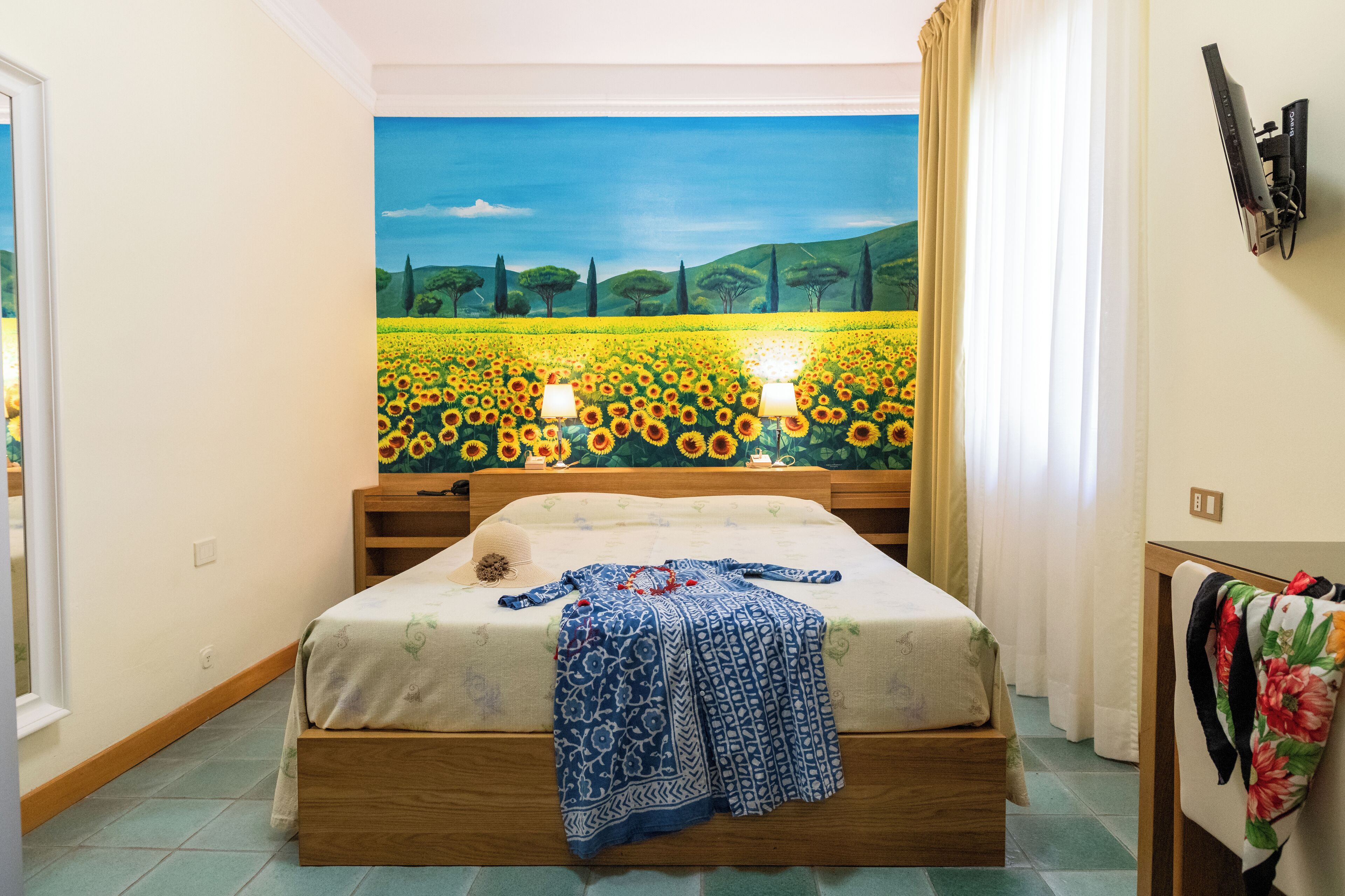 Ein Schlafzimmer mit einem großen Bett, einem Nachttisch, einer Lampe, einem Bild von einem Feld und einem Fenster mit Vorhängen.