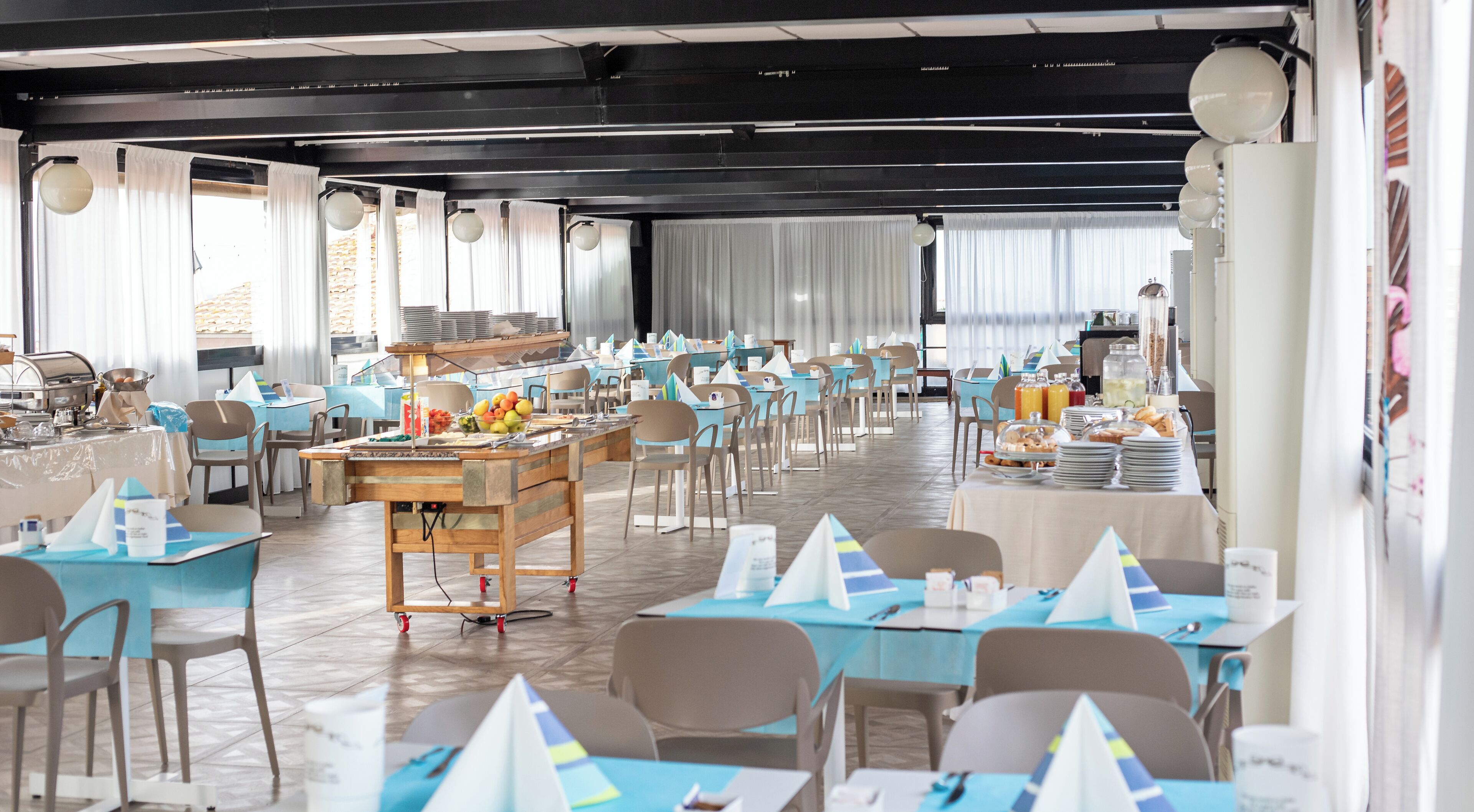 A spacious dining area with multiple tables and chairs, a buffet setup, and large windows allowing natural light.