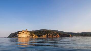 A coastal landscape with a rocky island featuring a small building, calm water, and distant hills.