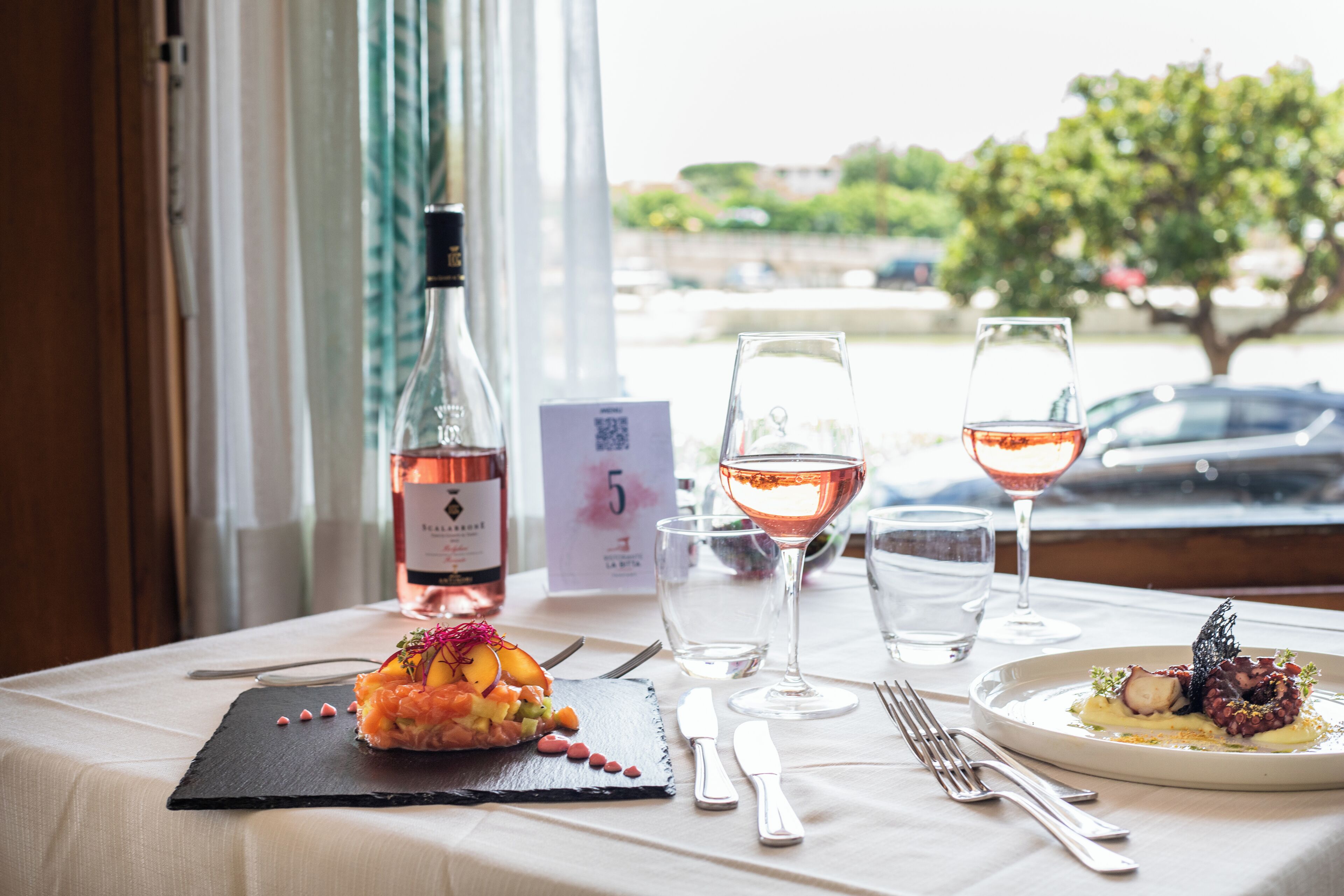 A table set with a meal, wine glasses, and a bottle of wine, with a view of a tree and a car in the background.