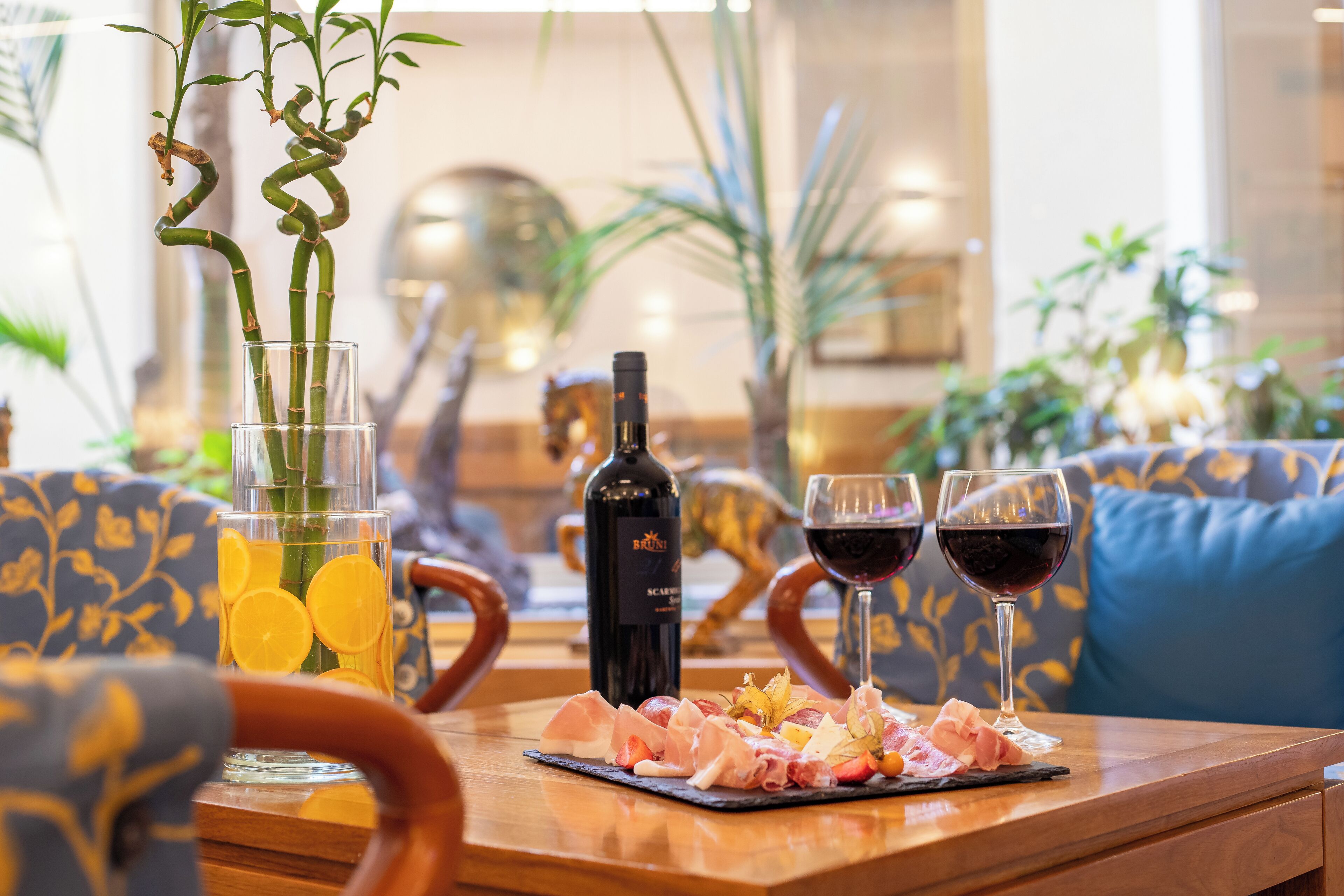 Une table à manger avec une bouteille de vin, deux verres de vin rouge, un verre d’eau avec des rondelles de citron et une assiette de nourriture.