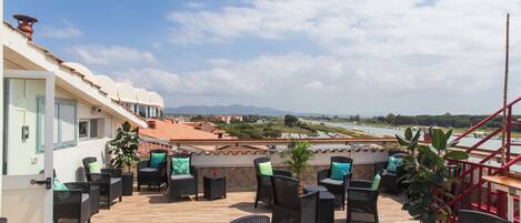 Terraza en la azotea con mobiliario de mimbre y plantas en macetas, con vistas a un paisaje urbano y una masa de agua.