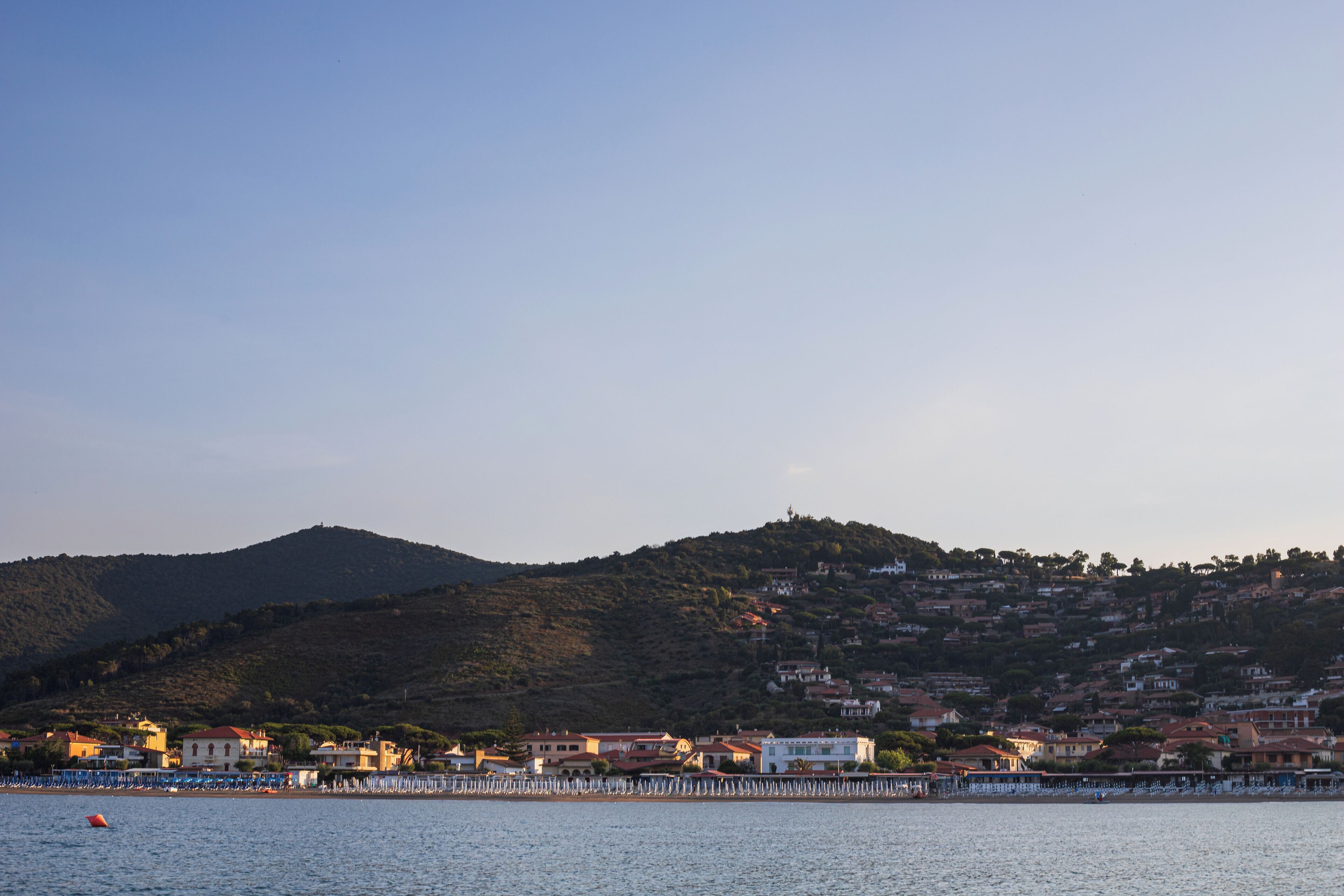 Un pueblo costero con edificios a lo largo del paseo marítimo, un muelle y una colina al fondo.