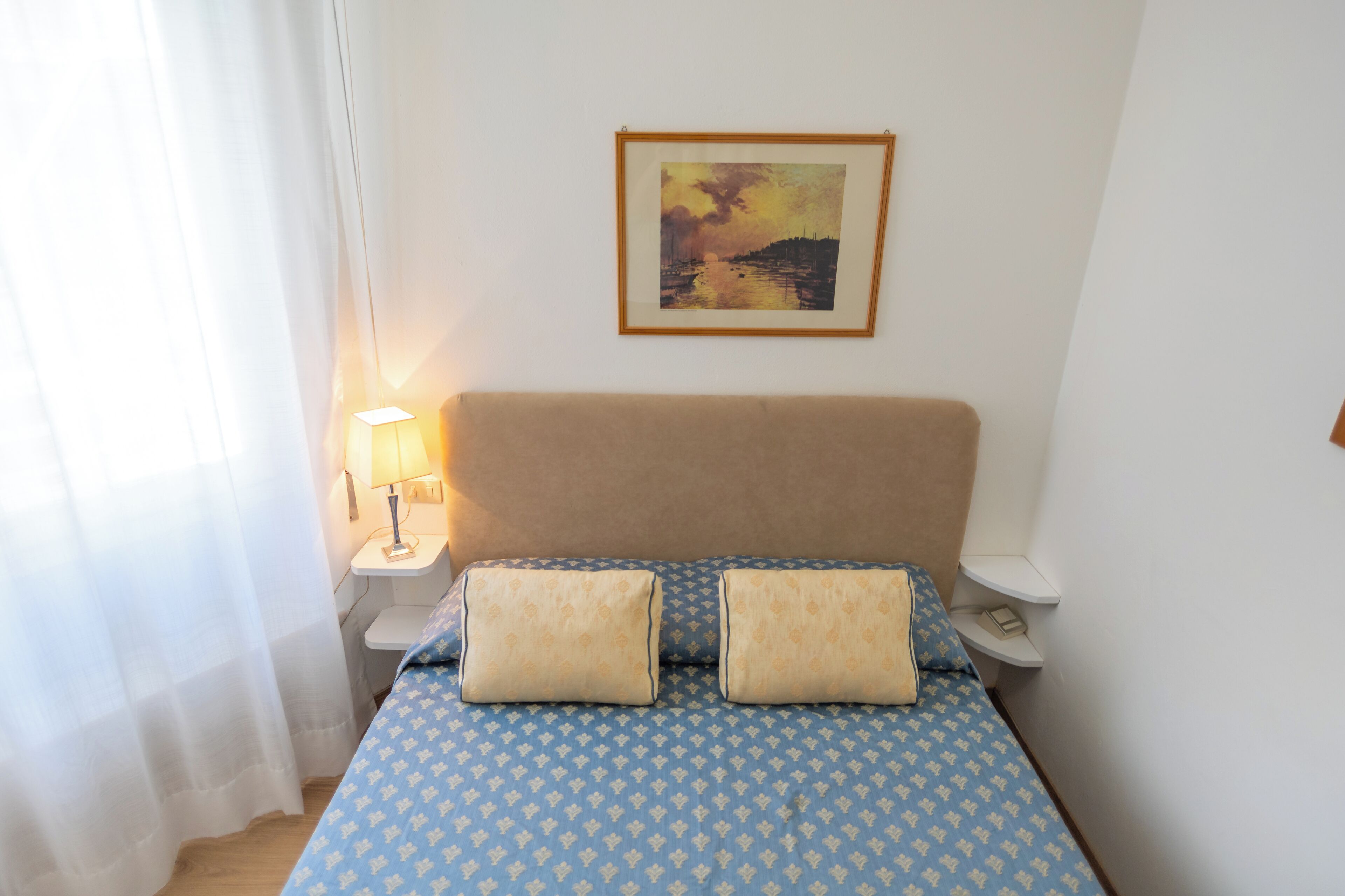 Un dormitorio con cama, dos lámparas de noche, mesita de noche, un cuadro en la pared y una ventana con cortinas.
