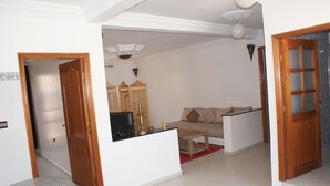 Una sala de estar con un sofá, una mesa de centro y un televisor. Se observa un aparador de madera y una mesita con una lámpara.