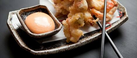 Un plato de camarones tempura con salsa para mojar y un tazón pequeño de salsa, acompañado de palillos sobre una mesa oscura.