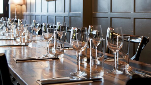 A long dining table with multiple wine glasses and cutlery, set against a wall with framed artwork and a wooden cabinet. - The One Tun Pub and Rooms (London)