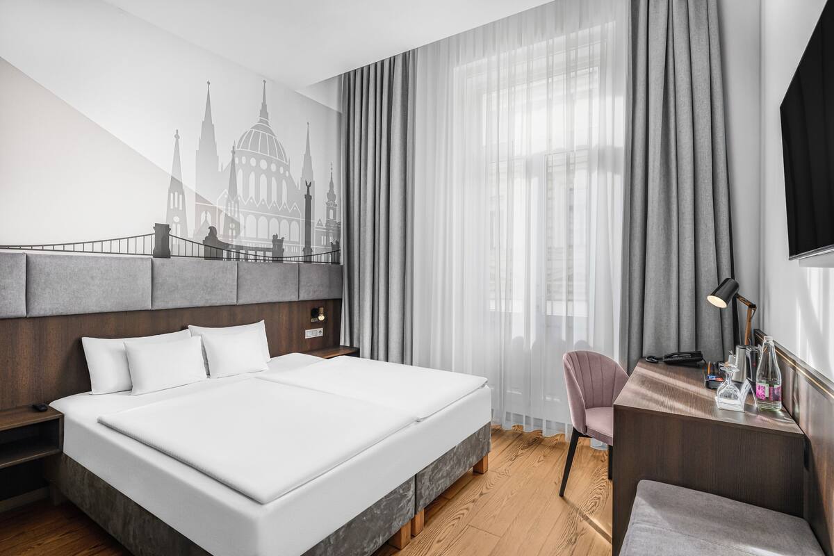 A hotel room with a large bed, a desk with a chair, a TV, and a wall mural of a building.
