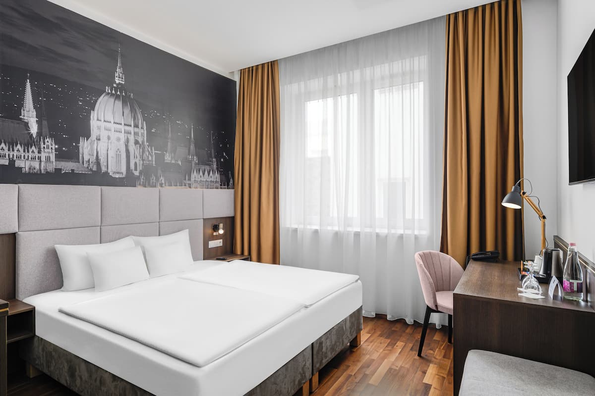 A hotel room with a large bed, a desk, a chair, and a cityscape mural on the wall.