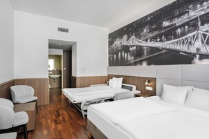 Habitación de hotel con dos camas, zona de estar, baño y un gran mural de la ciudad en la pared.
