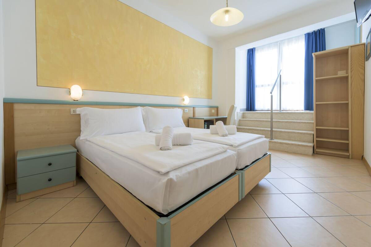A double bed with two pillows and two towels, a nightstand, a desk, a bookshelf, and a large window with curtains.