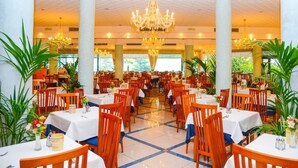 Uma sala de jantar espaçosa com várias mesas e cadeiras, grandes lustres e plantas em vasos.
