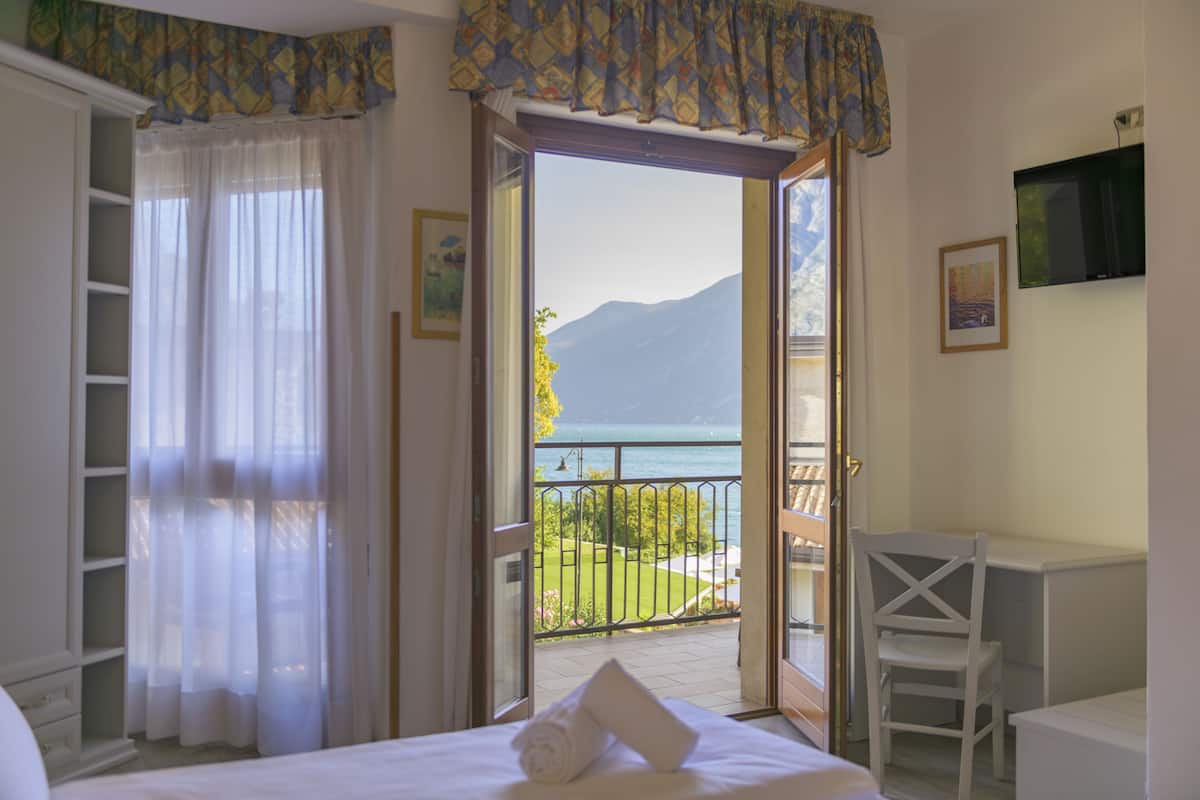 A bedroom with a bed, a chair, a desk, and a balcony with a view of the sea and mountains.