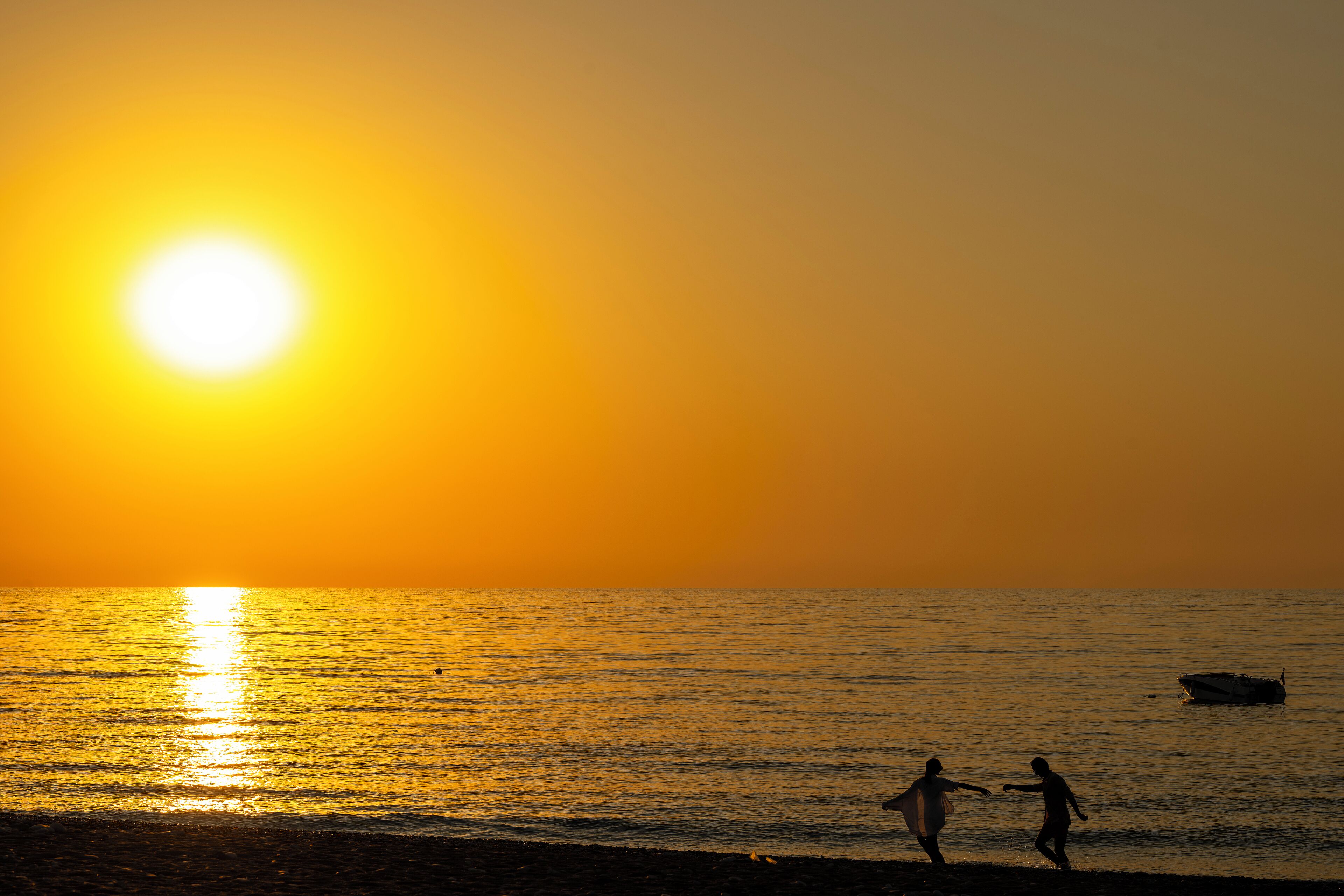 Ein Sonnenuntergang über einem ruhigen Meer, im Vordergrund die Silhouetten zweier Personen am Strand.