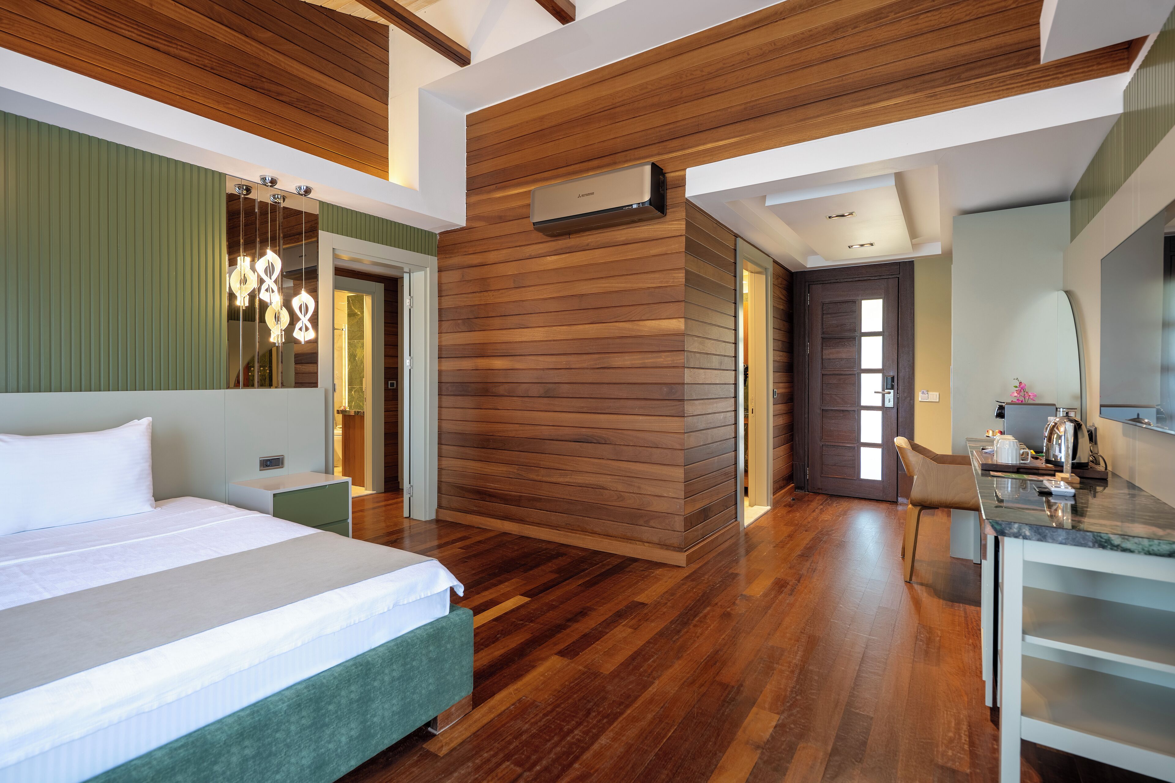 Habitación de hotel moderna con piso de madera, cama con ropa blanca, pared de acento verde, techo de madera y zona de cocina con encimera de mármol.